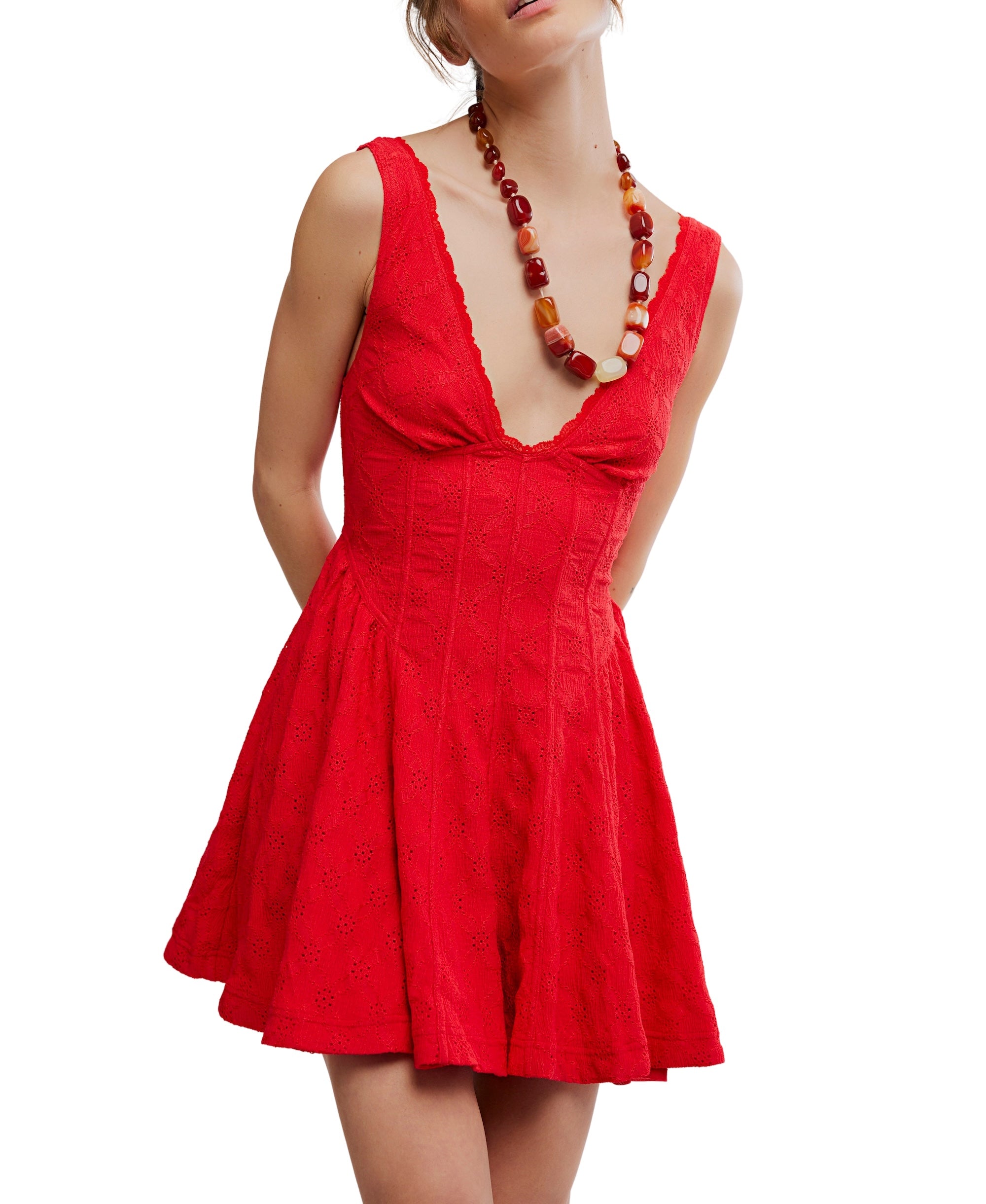 A woman models the Free People Cant Help It Mini, a sleeveless red dress with a deep V-neck and A-line skirt, accessorized with a chunky beaded necklace, posing with her hands behind her back against a white background.
