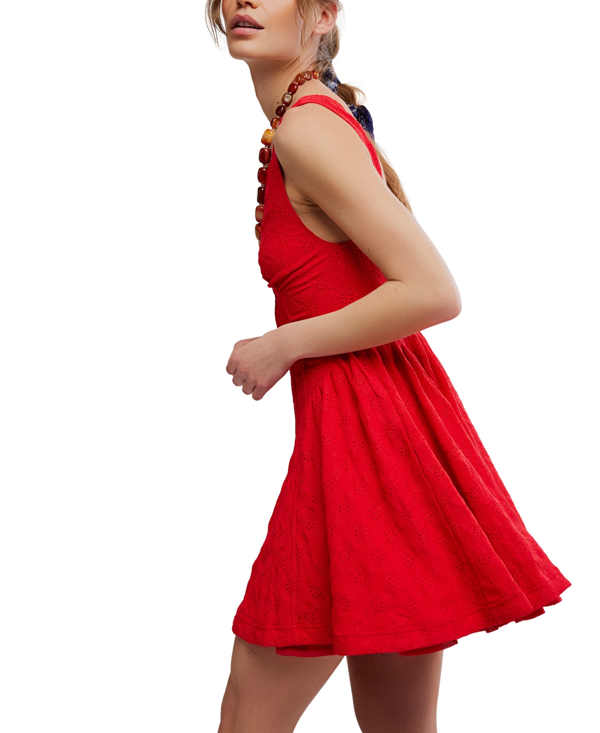 A woman models the Cant Help It Mini by Free People—a sleeveless, knee-length red dress with a corset-style bodice—standing sideways against a white background with her hair pulled back and wearing a beaded necklace.