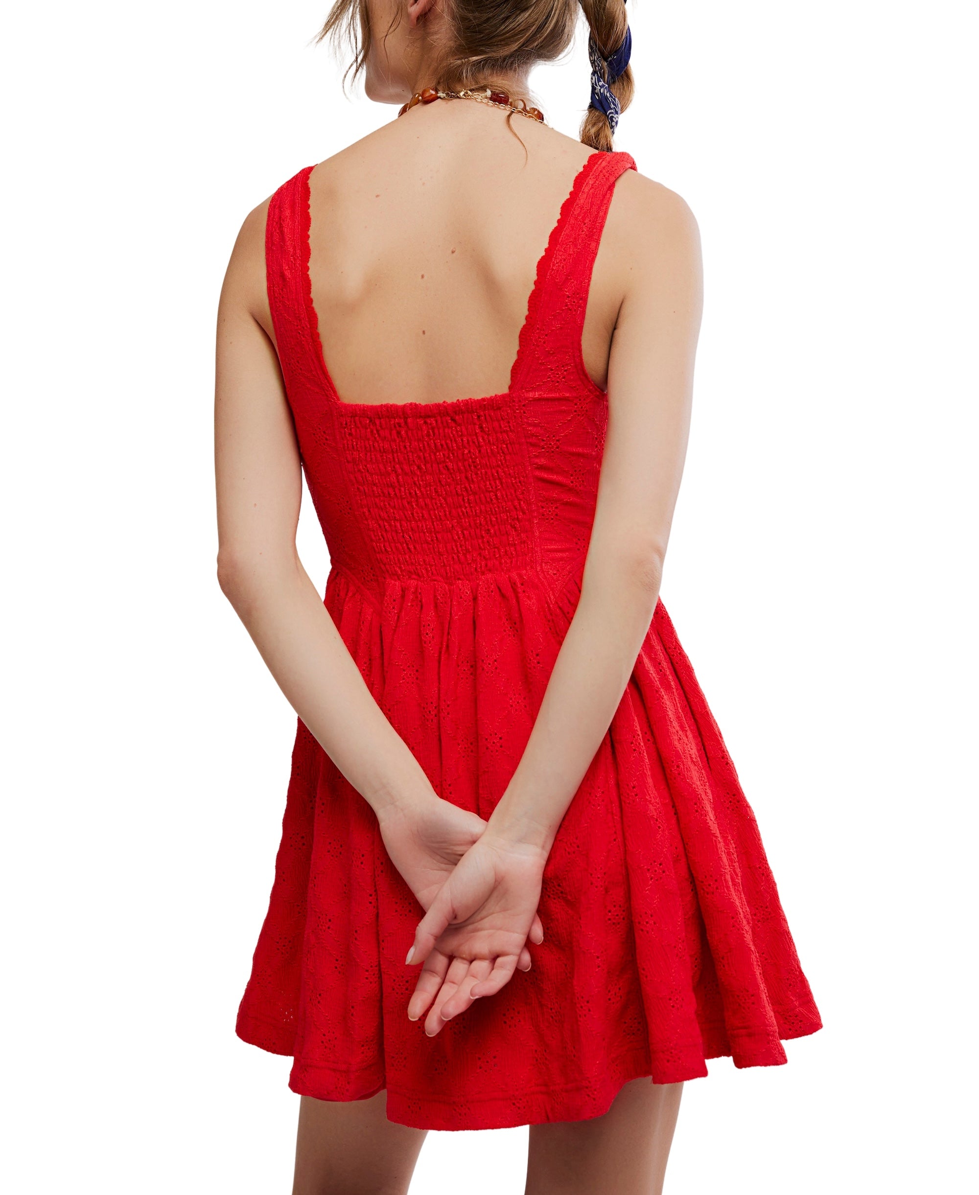 A woman with a braid wears the Free People Can’t Help It Mini—a sleeveless, bright red dress with a corset-style bodice and flared A-line skirt. She stands with her back to the camera and her hands clasped behind her.