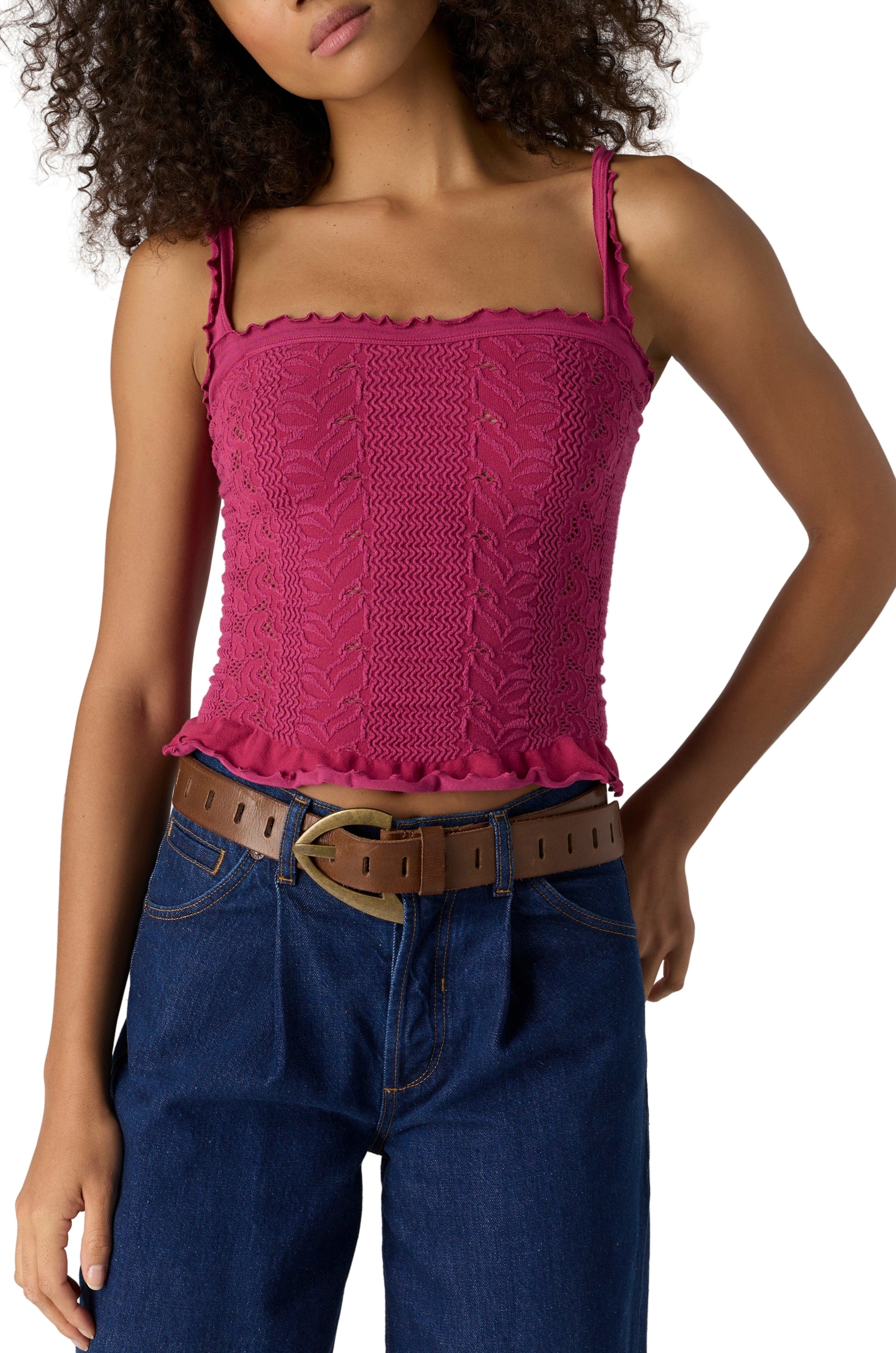 A woman with curly hair wears the Free People So Soft Femme Square Neck top in SANGRIA, featuring ruffle trim, paired with blue jeans and a brown belt with gold buckle. Her hand rests on her hip.