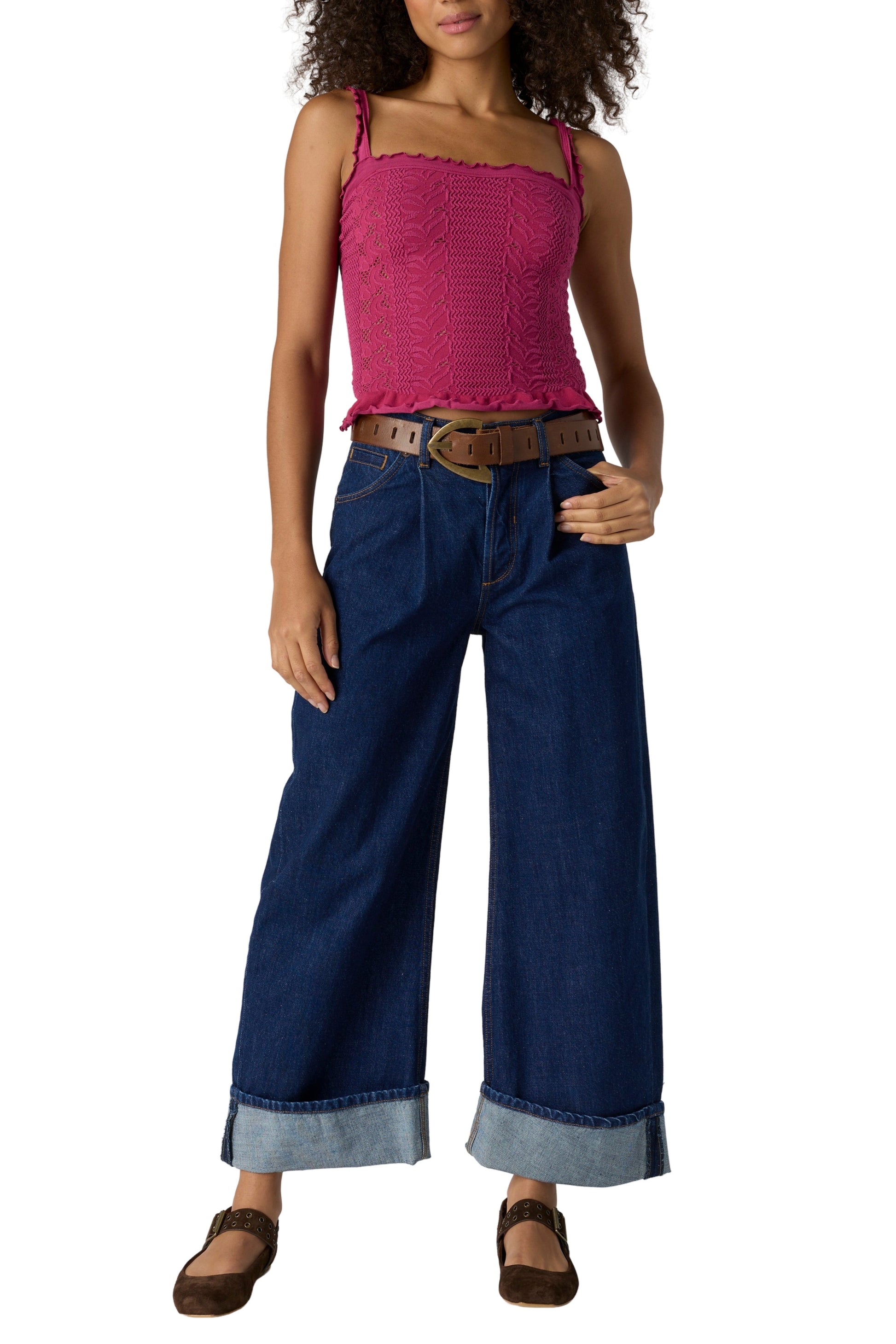 A woman wears the Free People So Soft Femme Square Neck top in magenta, wide-leg cuffed blue jeans, a brown belt, and brown shoes, posing with one hand on her hip against a white background.
