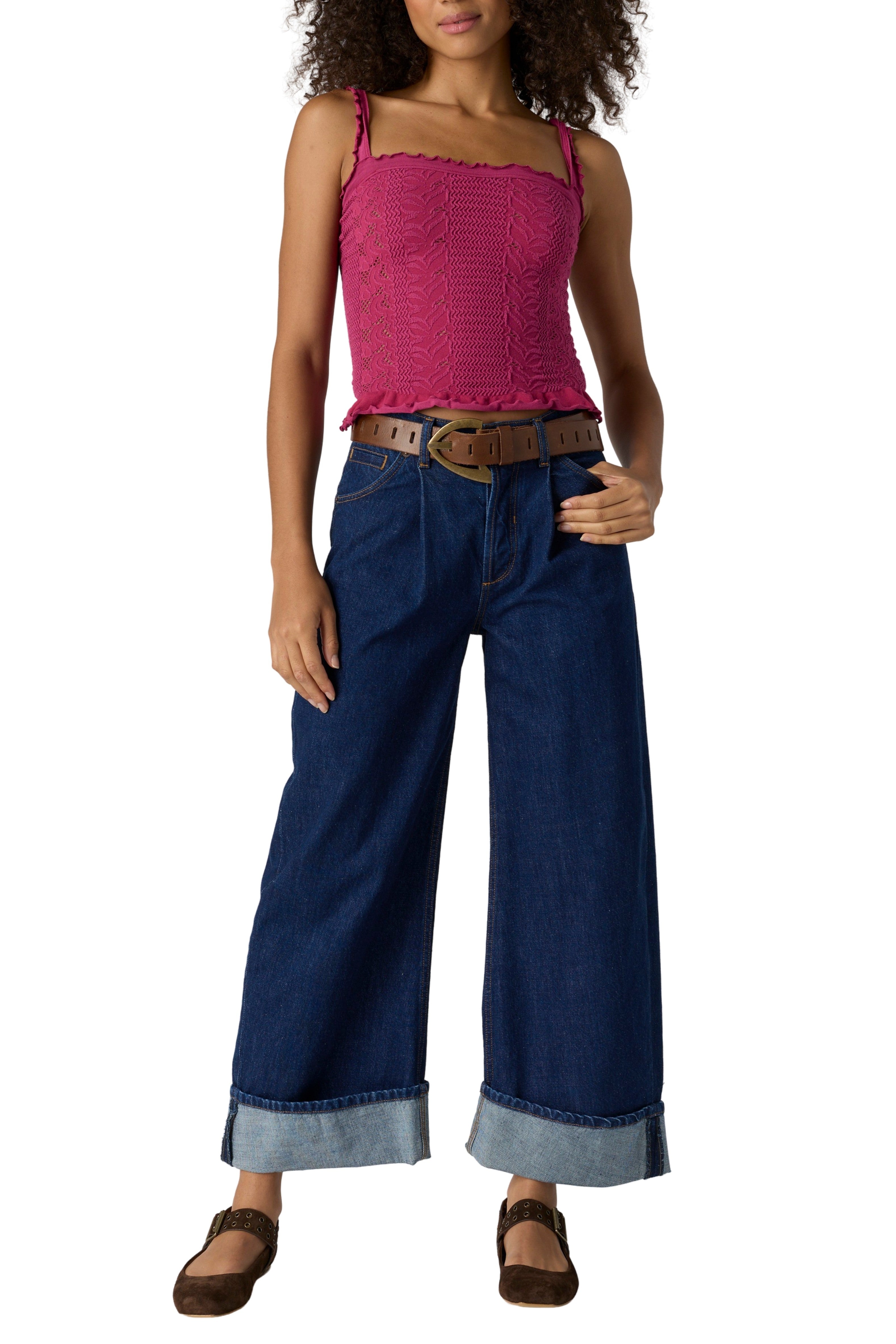 A woman wears the Free People So Soft Femme Square Neck top in magenta, wide-leg cuffed blue jeans, a brown belt, and brown shoes, posing with one hand on her hip against a white background.