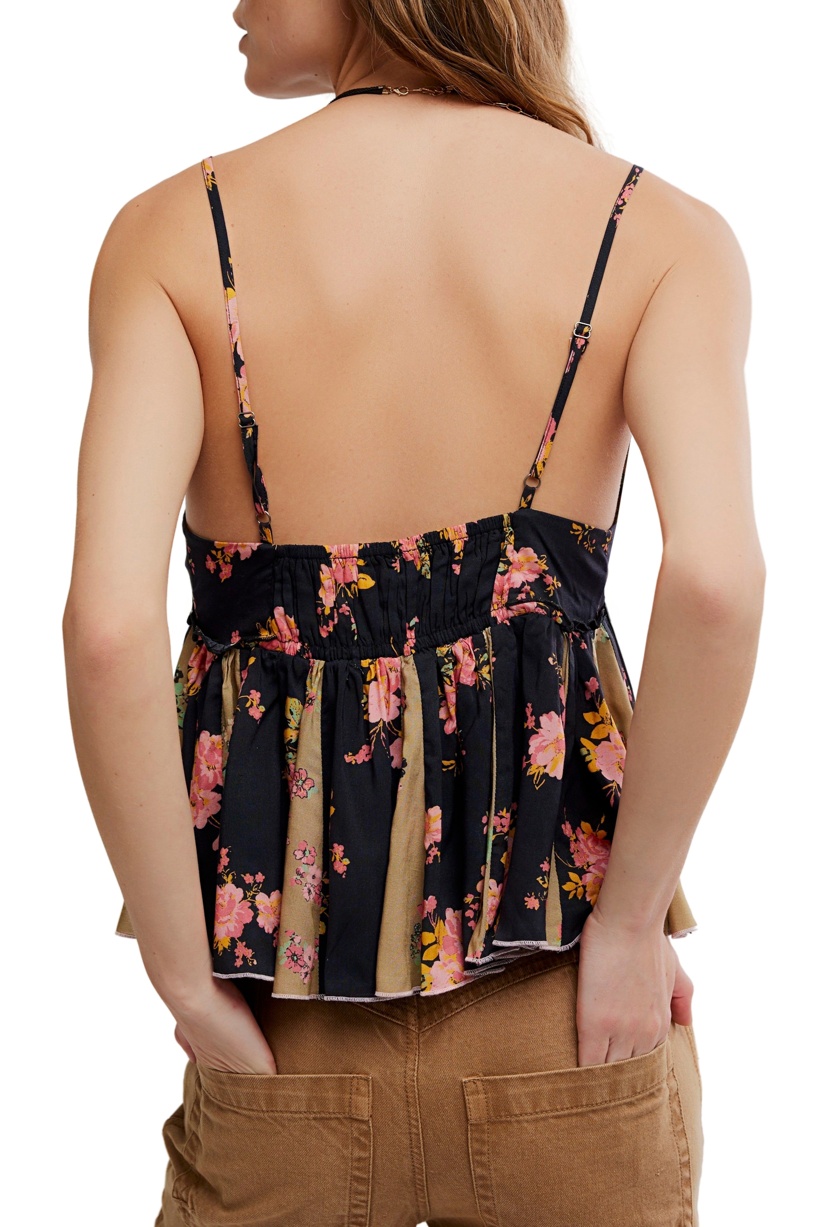 A woman is shown from the back wearing the Free People Tillie Top, a black floral print, backless camisole with adjustable straps, paired with brown pants. Her long hair is down and her hands are in her pockets.