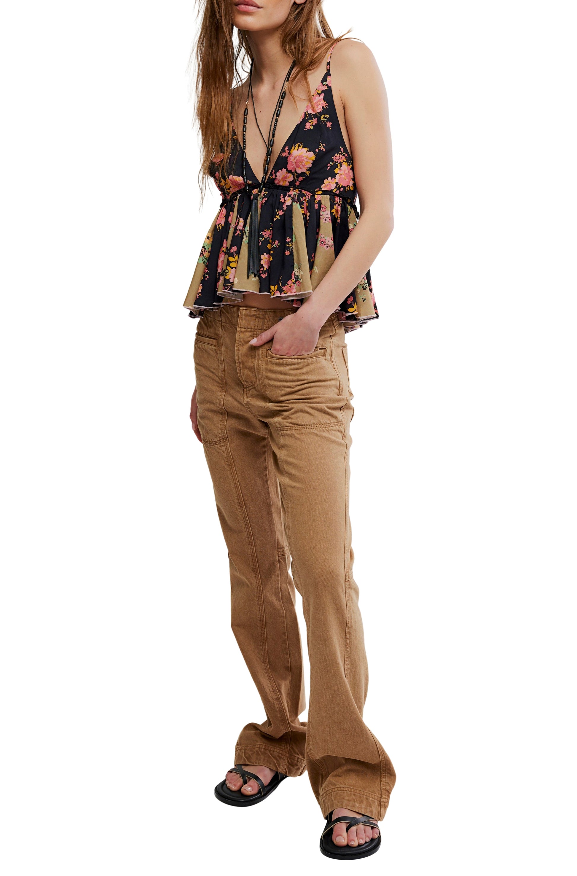 Wearing the Free People Tillie Top—a sleeveless, dark floral peplum with adjustable straps—paired with light brown high-waisted pants and black sandals, a person stands with one hand in their pocket against a white background.