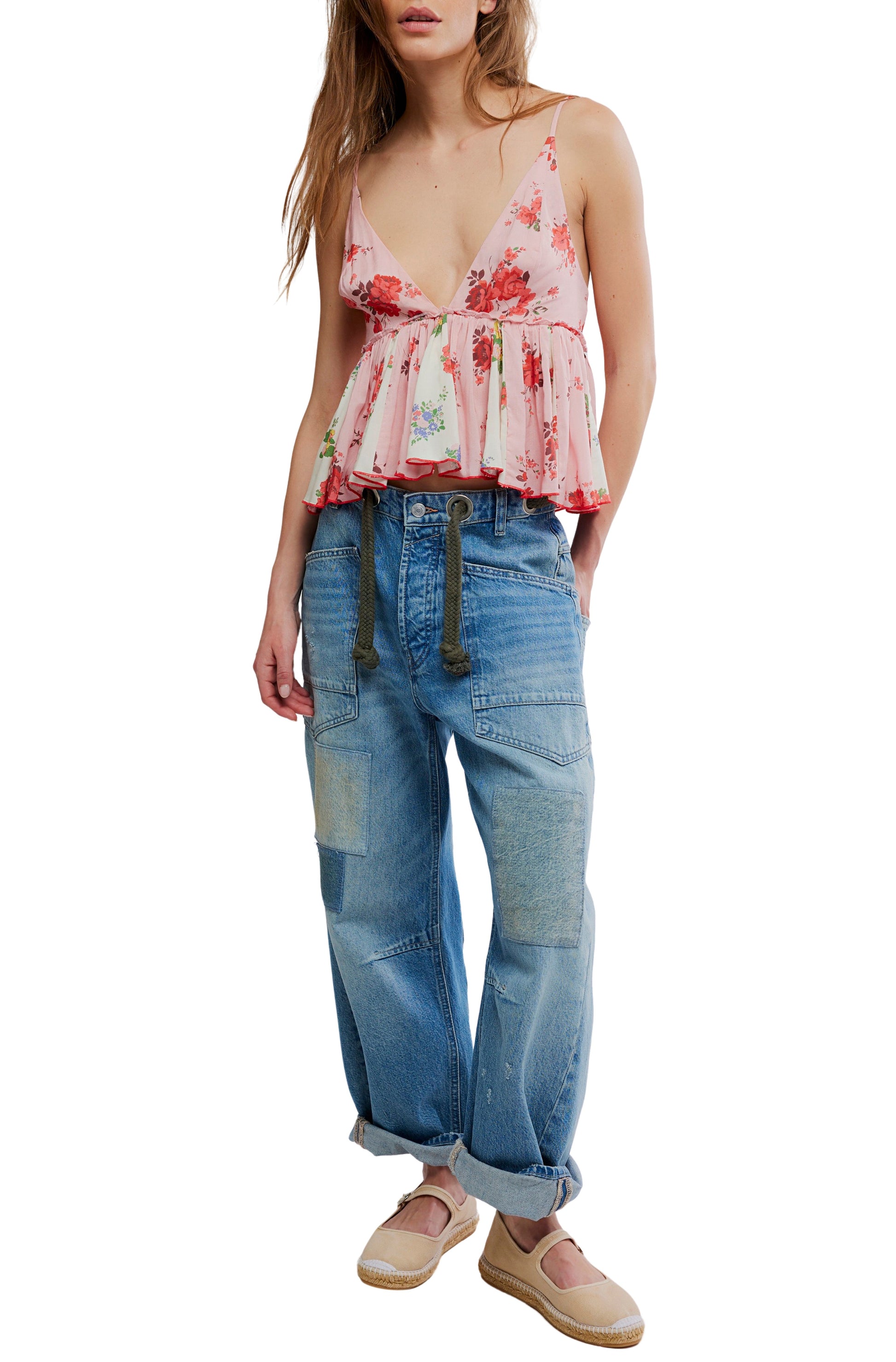 A person wearing the Free People Tillie Top—a pink floral sleeveless peplum with adjustable straps—paired with loose patchwork blue jeans, a rope belt, and beige espadrille flats, stands with hands in pockets.