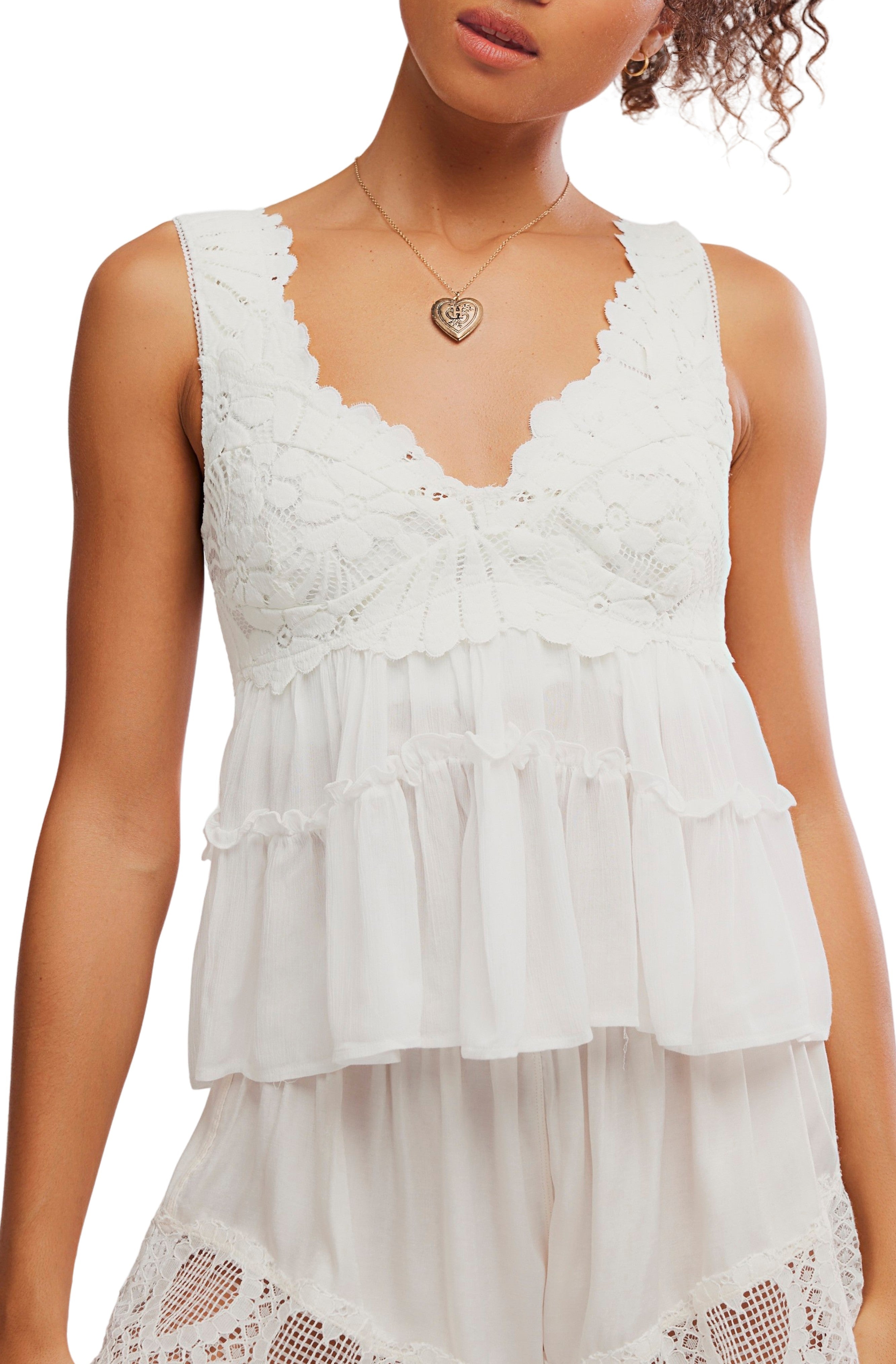 A woman wears the Free People Petal Party Shapeless top in ivory, featuring sleeveless lace and ruffles with a sweetheart neckline, paired with matching ivory shorts. She accessorizes with a heart-shaped pendant necklace and has curly hair.