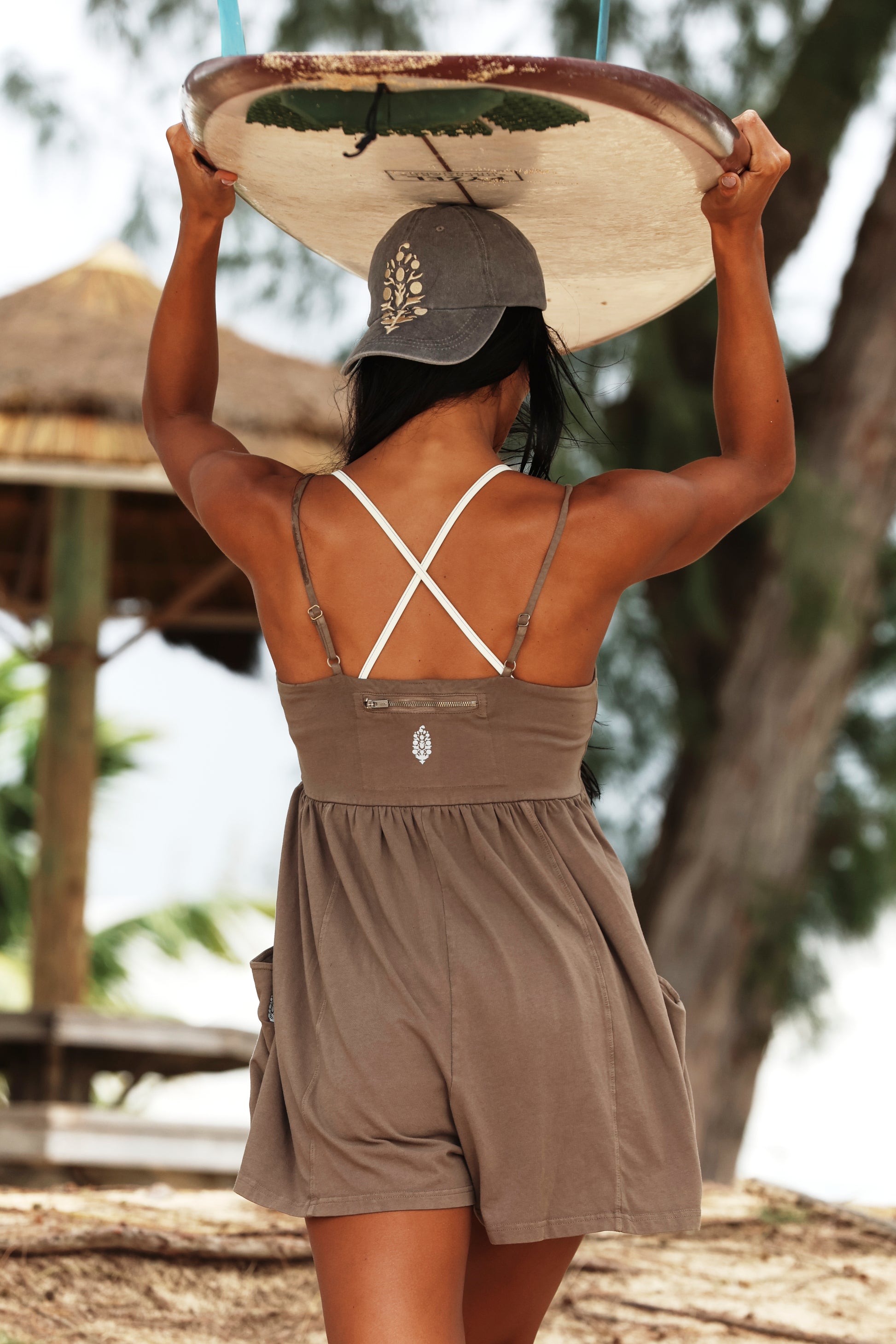 A woman with long dark hair, wearing the Hot Shot Strappy Romper by FP Movement, holds a surfboard above her head while walking outdoors near trees and a thatched-roof structure.