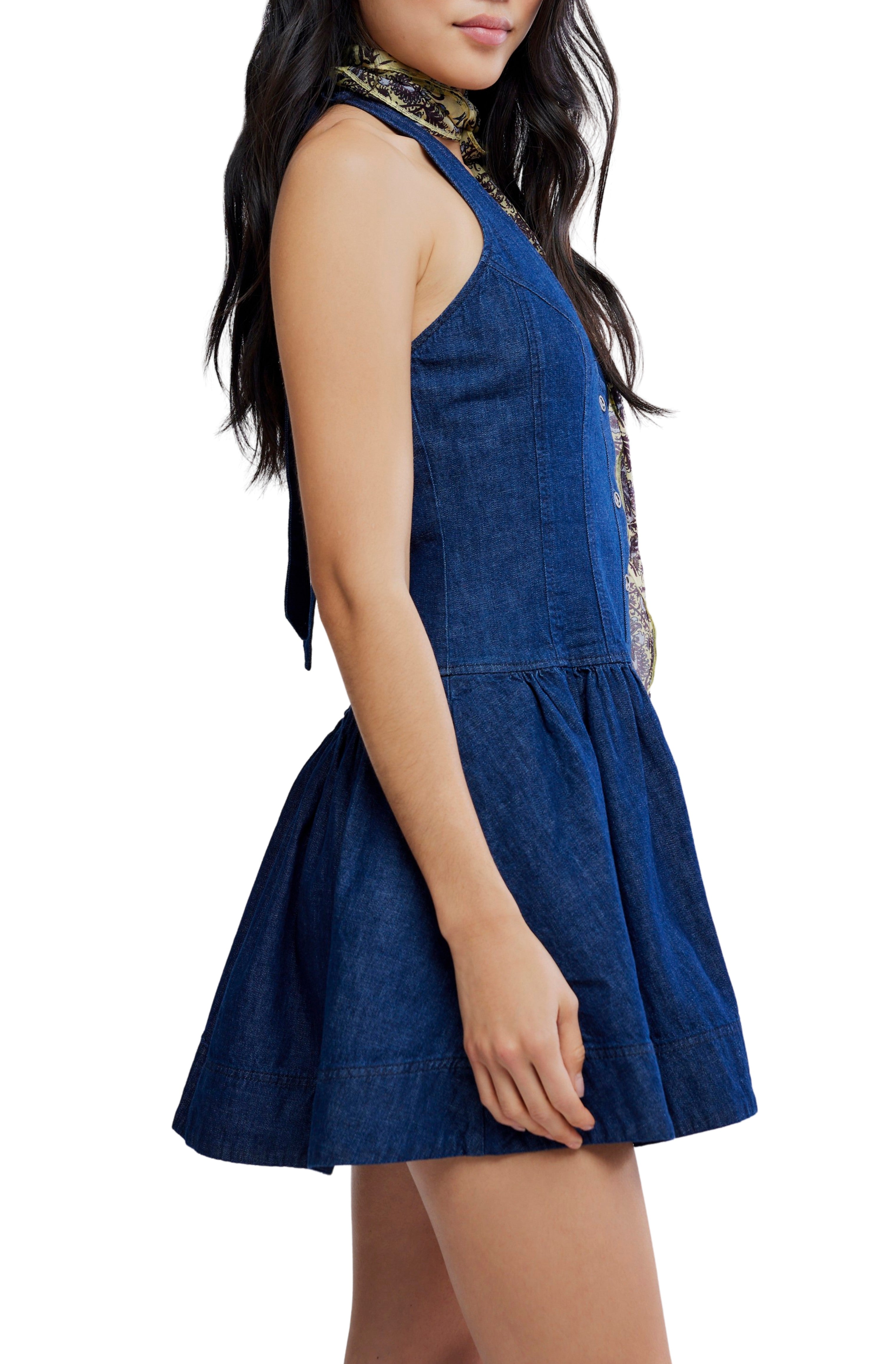 A woman stands sideways in the Clean Break Denim Mini Dress by Free People, a sleeveless dark blue denim piece with a flared skirt and button-front closure. She wears her long dark hair loose and has a patterned scarf around her neck.