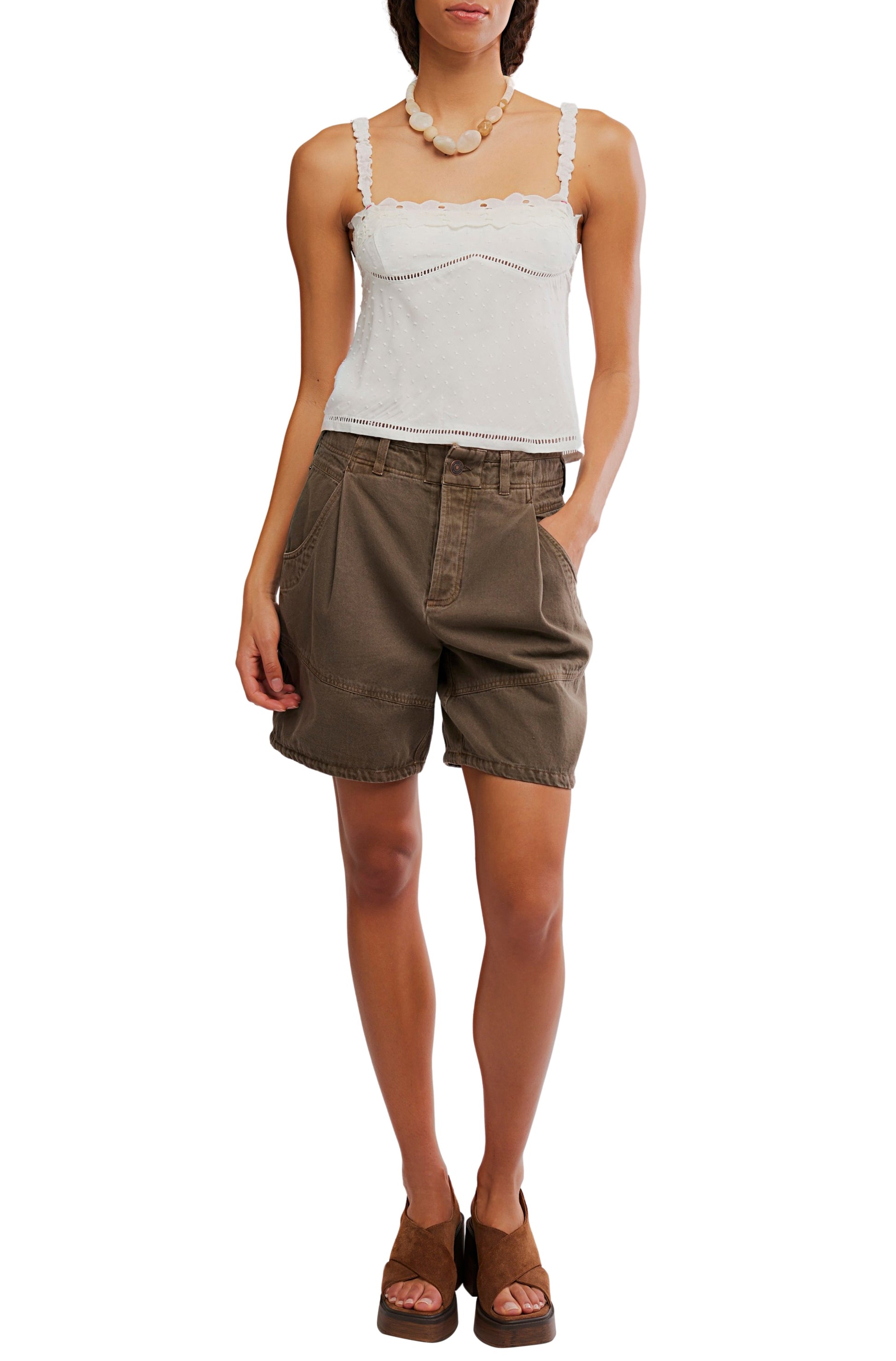 Wearing the Free People Little Love Tank in white with adjustable straps, loose brown pocket shorts, brown wedge sandals, and a chunky beaded necklace, a person stands casually with one hand in their pocket.