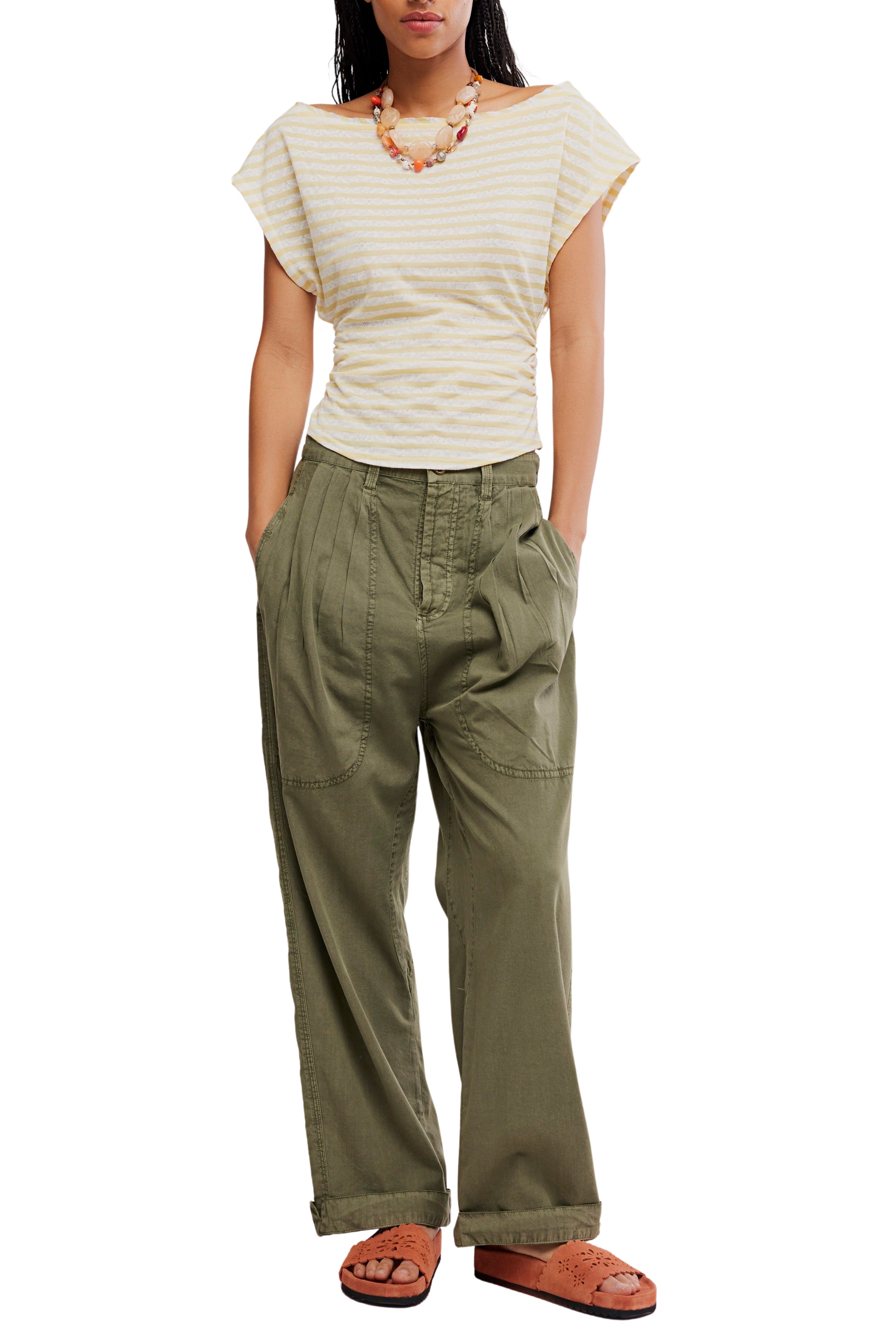 A person wearing the Free People Ellie Top—an ivory striped off-the-shoulder short-sleeve style—paired with olive wide-leg pants, brown sandals, and a chunky necklace stands with hands in pockets against a white background.