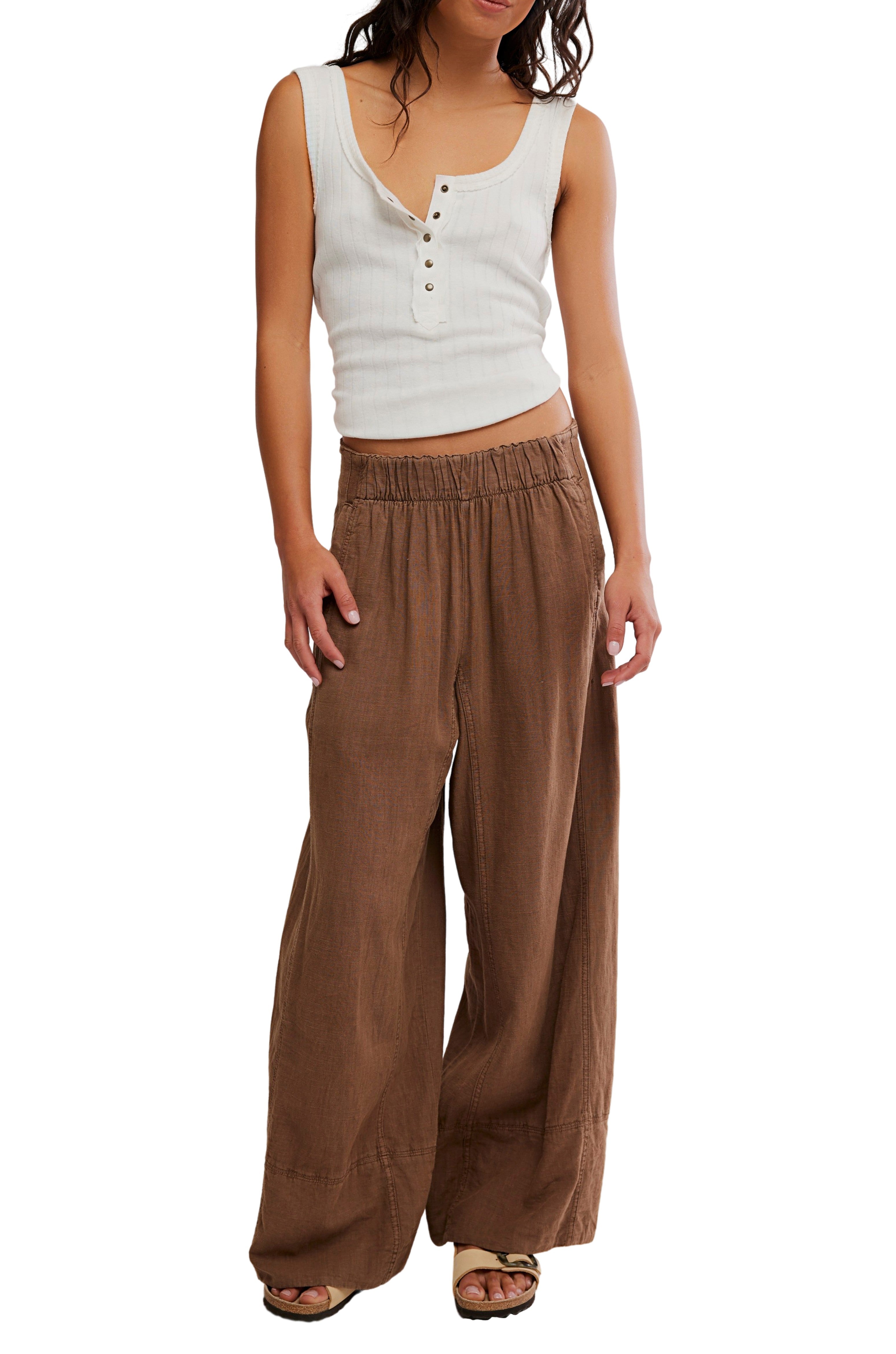 A person wears the Days End Linen Pull On Pant by Free People—loose, mid-rise, brown, wide-leg pants—paired with a white sleeveless button-front crop top, posing against a white background.