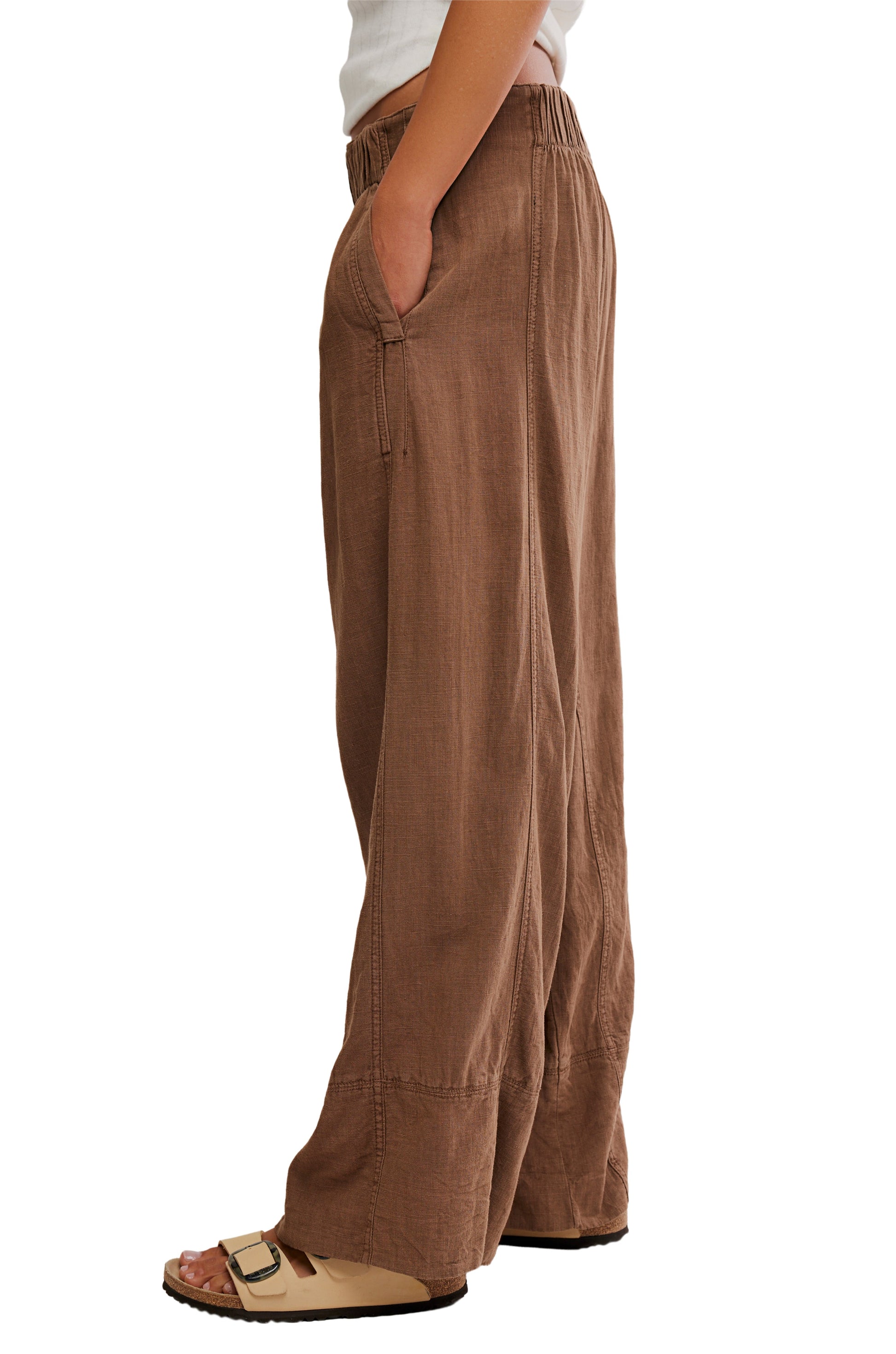 A person wearing the Free People Days End Linen Pull On Pant in brown with a natural texture and side pockets, paired with beige sandals, stands with one hand in a pocket against a white background.