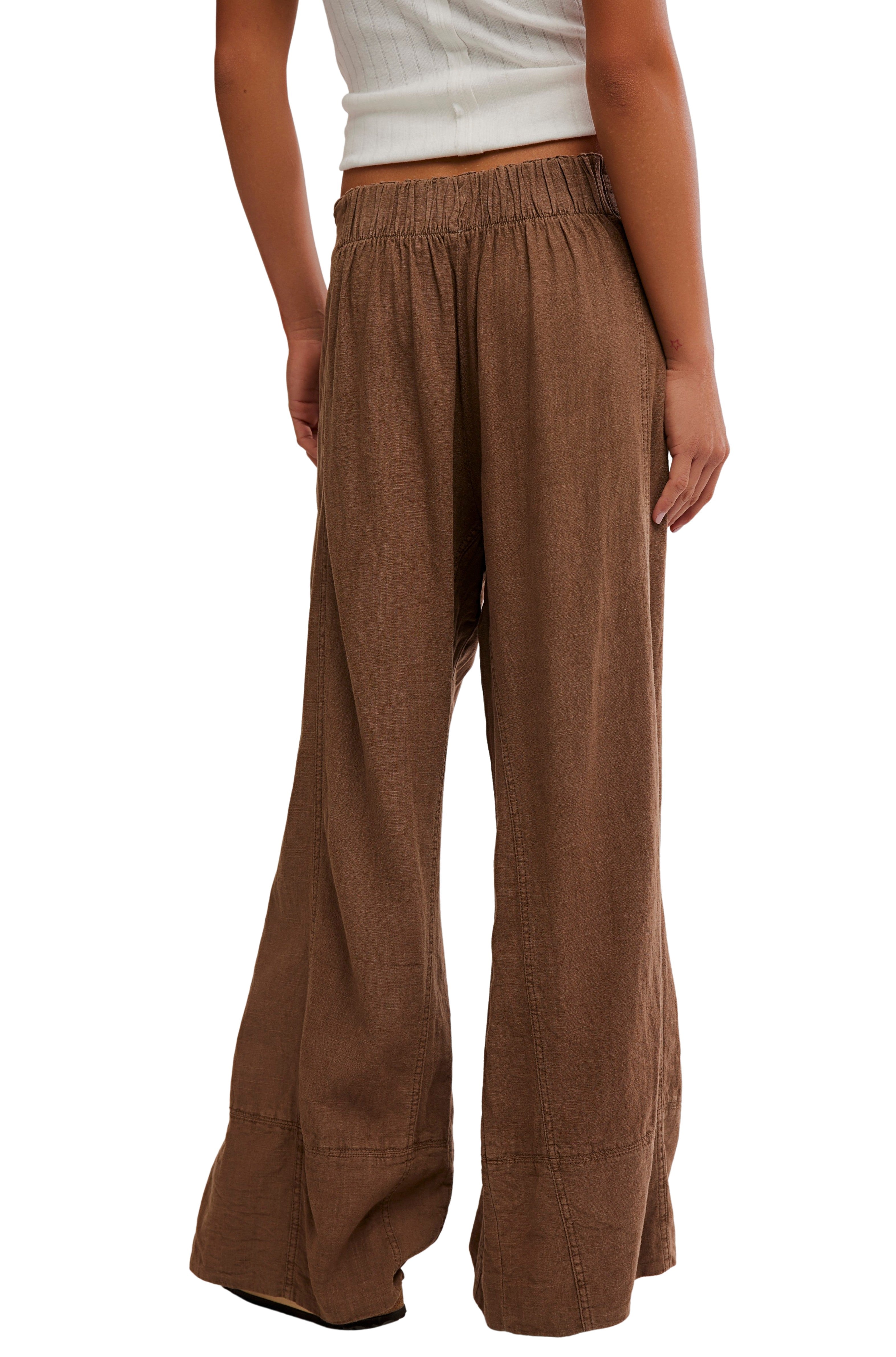 A person is seen from behind against a white background, wearing Free People's Days End Linen Pull On Pant—loose, wide-leg brown pants with a mid-rise elastic waistband and a natural texture—paired with a sleeveless white top.
