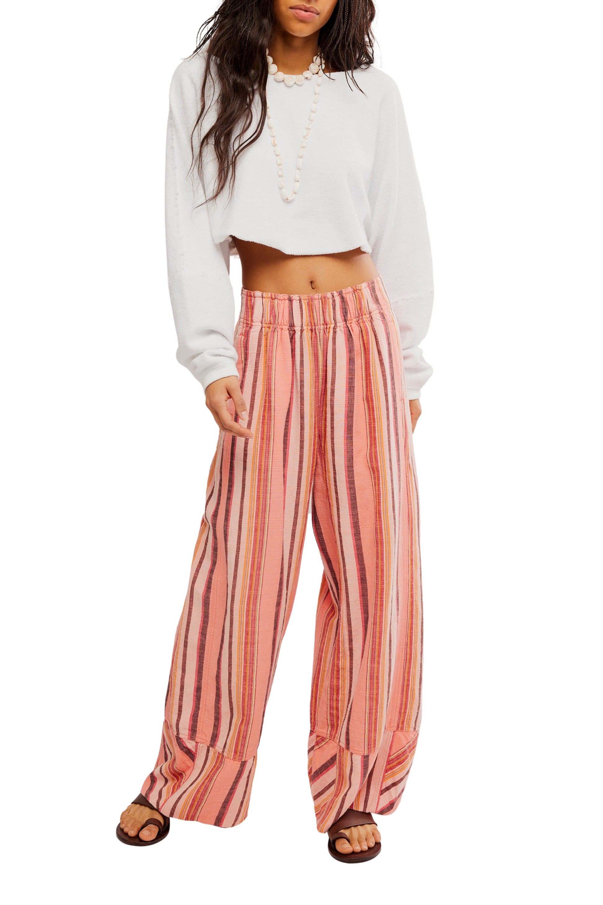 A person wearing Free People’s Days End Solar Stripe Pant—mid-rise, loose, pink and orange striped wide-leg pants—paired with a white long-sleeve cropped top and black sandals, stands against a white background.