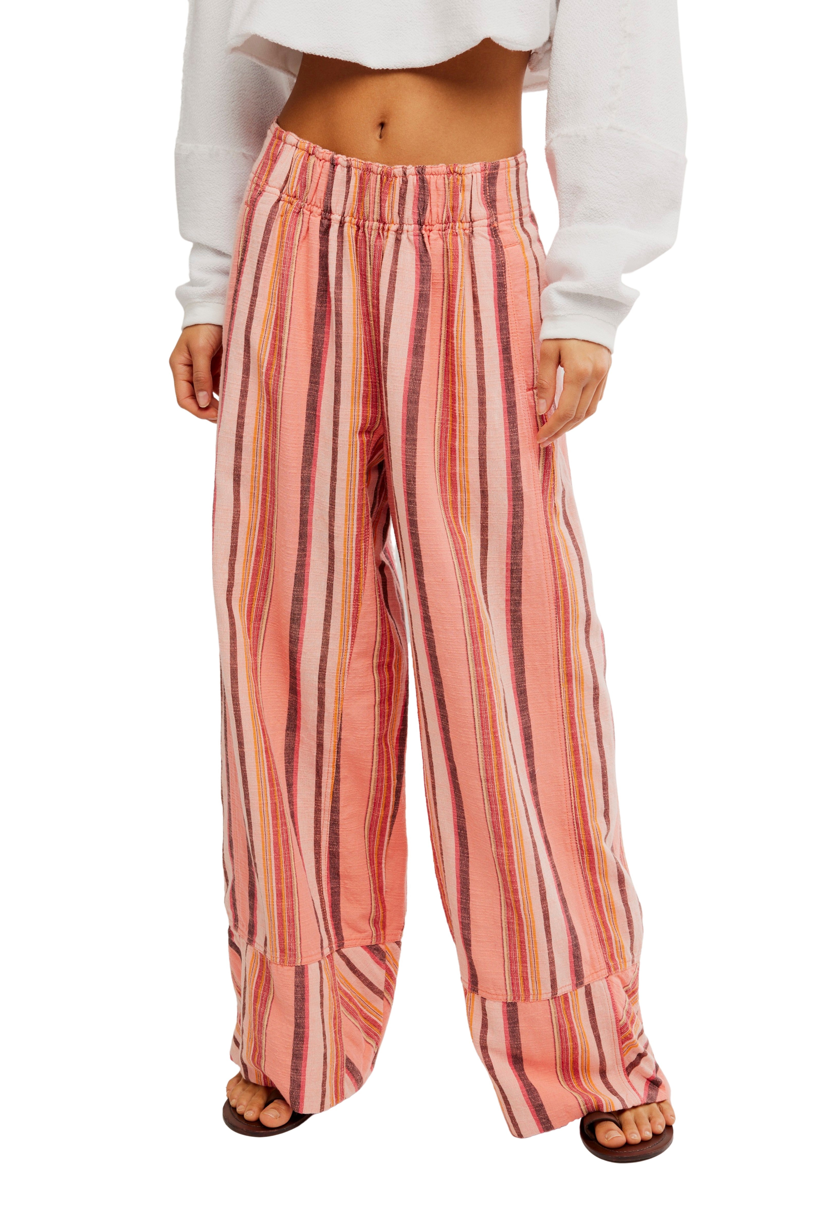 A person wears Free People's Days End Solar Stripe Pant in Soft Coral Combo, featuring loose, mid-rise cotton-linen stripes, paired with a white cropped long-sleeve top and sandals. The photo is cropped at the shoulders.