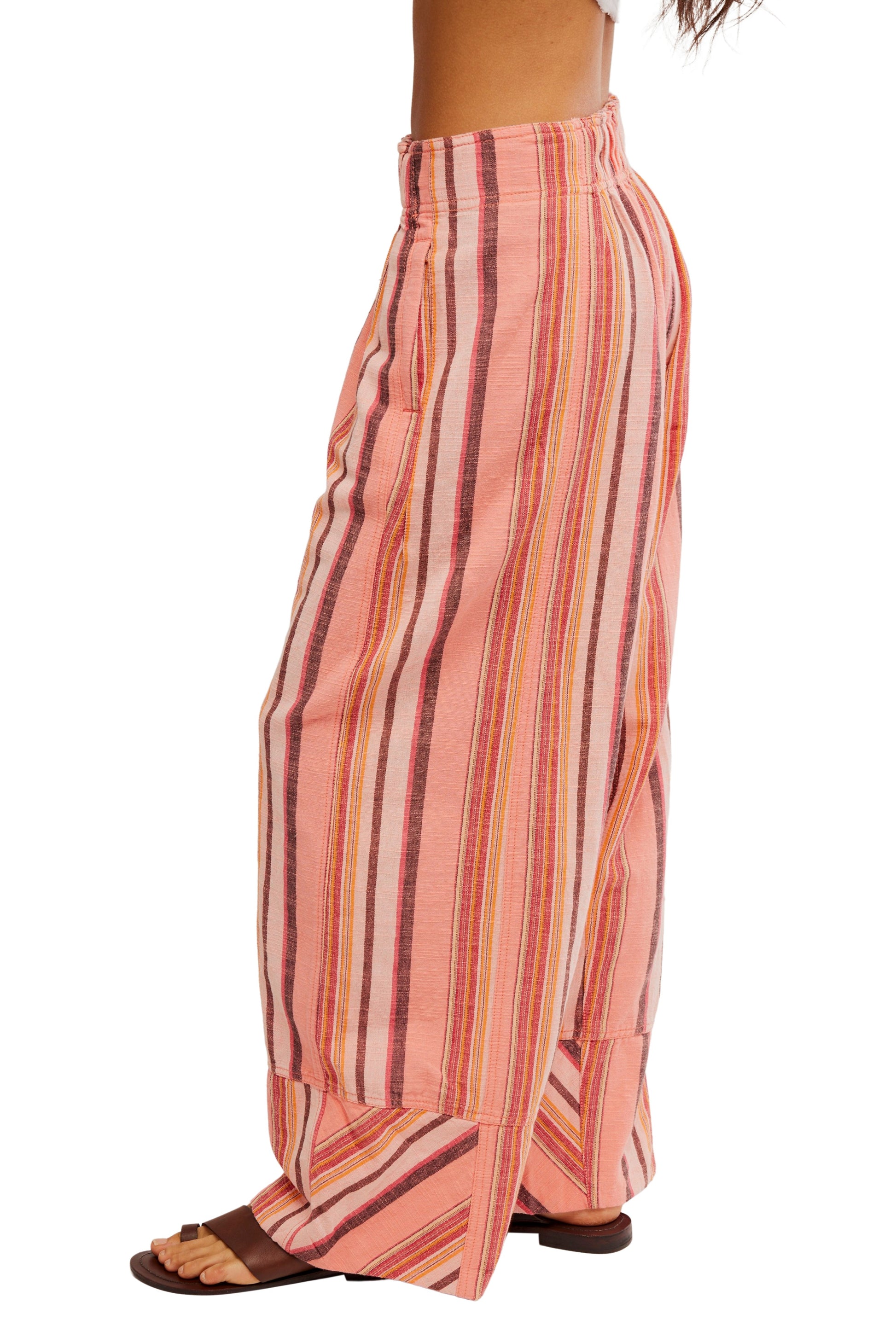 A person wearing Free People’s Days End Solar Stripe Pant—mid-rise, wide-leg with vertical pink, red, and white stripes in a natural textured finish—is shown from waist to feet in side view, paired with brown sandals.
