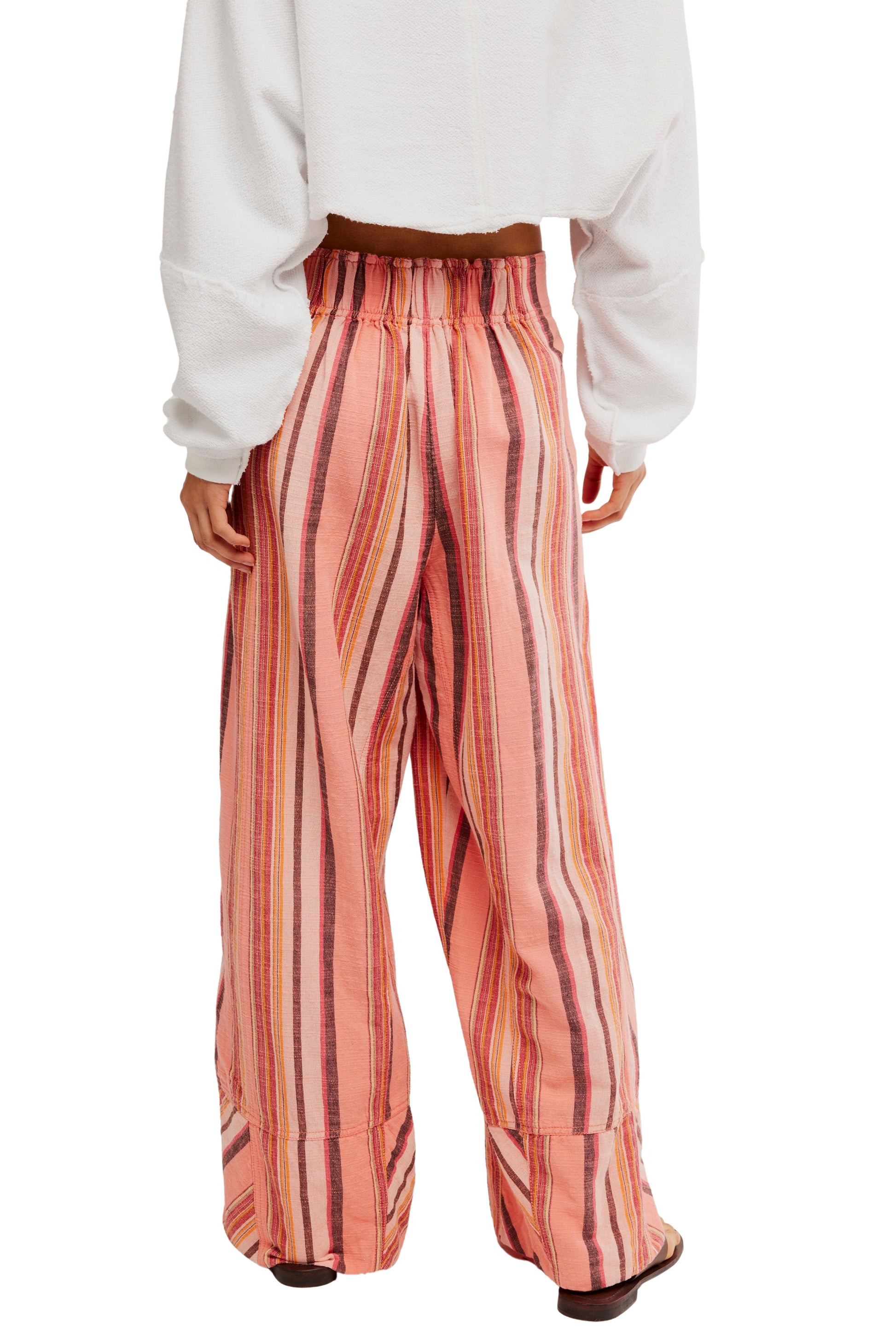 Model wearing the Free People Days End Solar Stripe Pant, featuring loose-fit, mid-rise, wide-leg style with vertical pink, orange, and brown stripes in a natural textured finish, paired with a cropped white long-sleeve top and sandals.