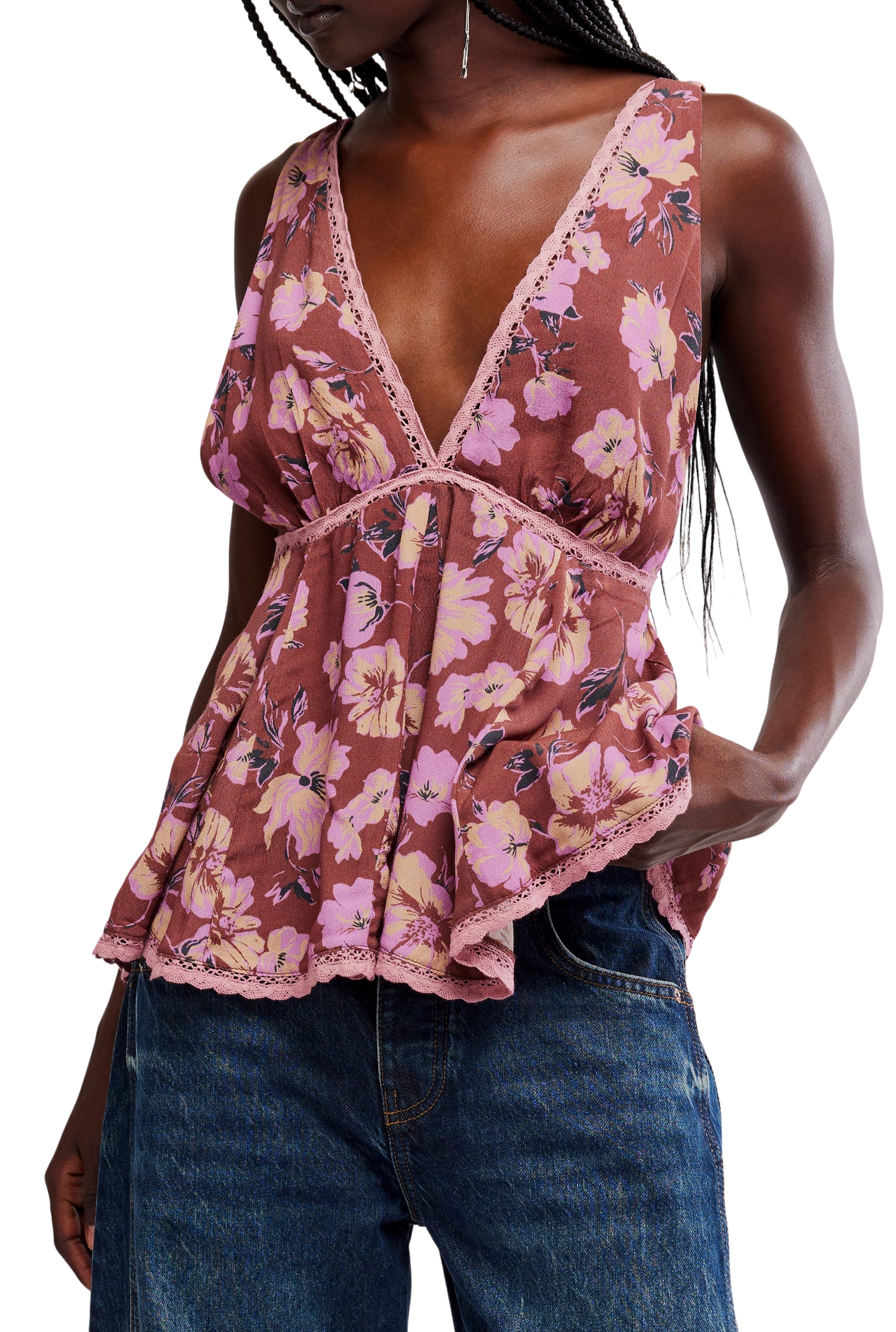 A person wears the Free People Rome Printed Tank in BROWN COMBO, featuring a sleeveless, deep V-neck floral design and lace trim, paired with blue denim jeans for a loose, flowy fit as they casually slip one hand in their pocket.