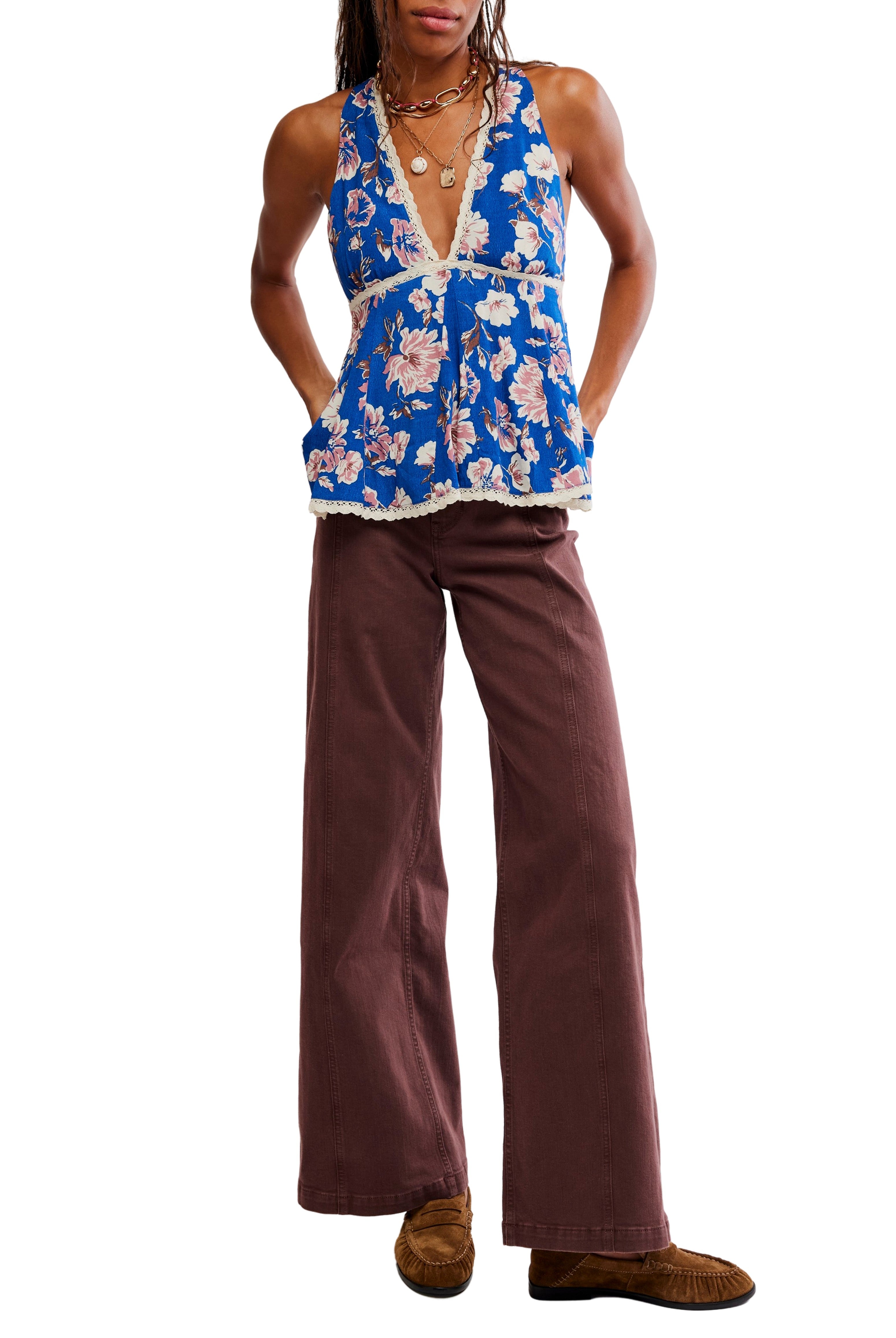 Model wears the Free People Rome Printed Tank—a blue floral sleeveless top with a plunging halter neckline and lace trim—paired with brown wide-leg pants, brown shoes, and layered necklaces. Made from 100% viscose.