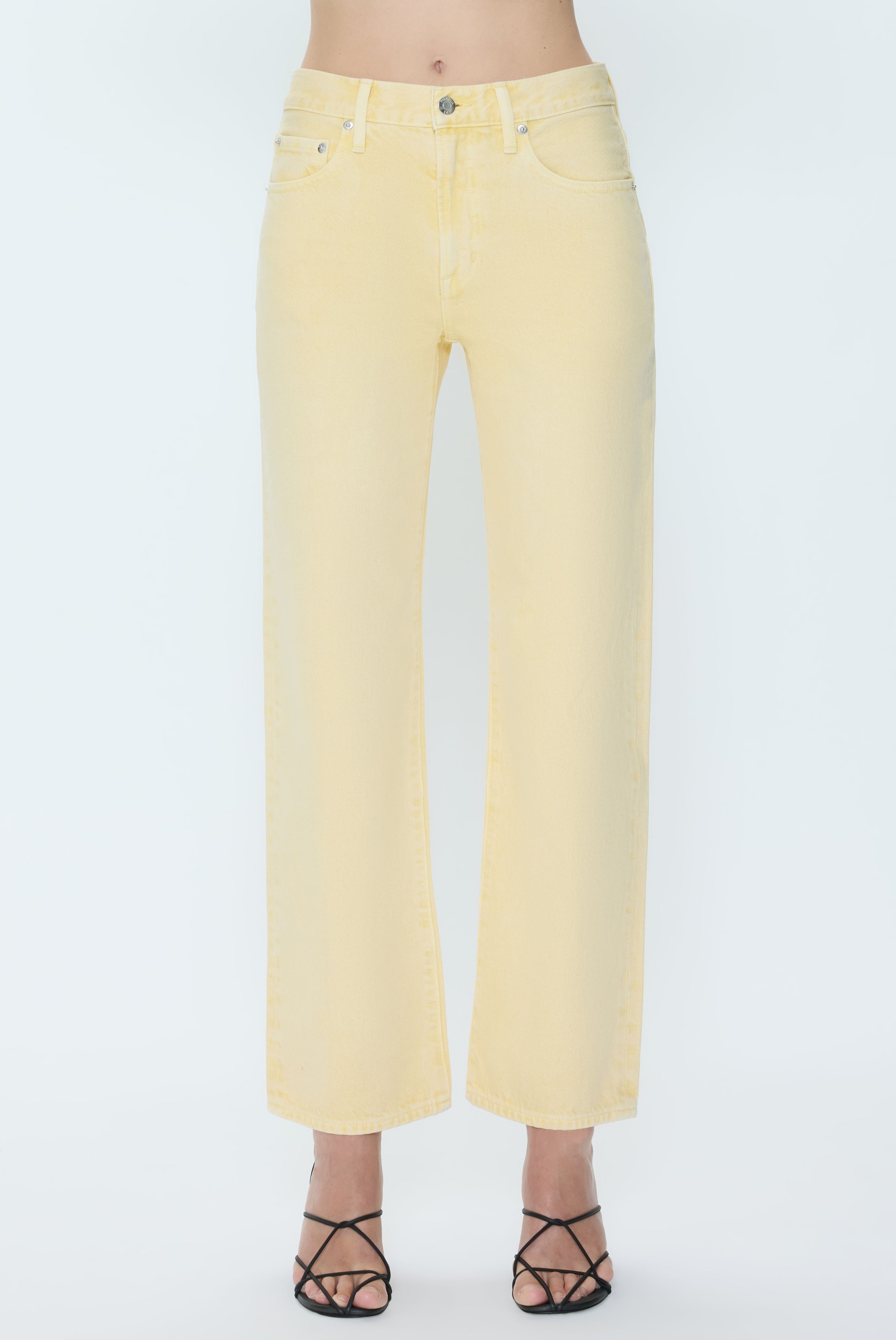 A person models the Pistola Lexi - Mid Rise Bowed Straight jeans in yellow, paired with black strappy heeled sandals, standing against a plain white background. Only the lower body is visible in the image.