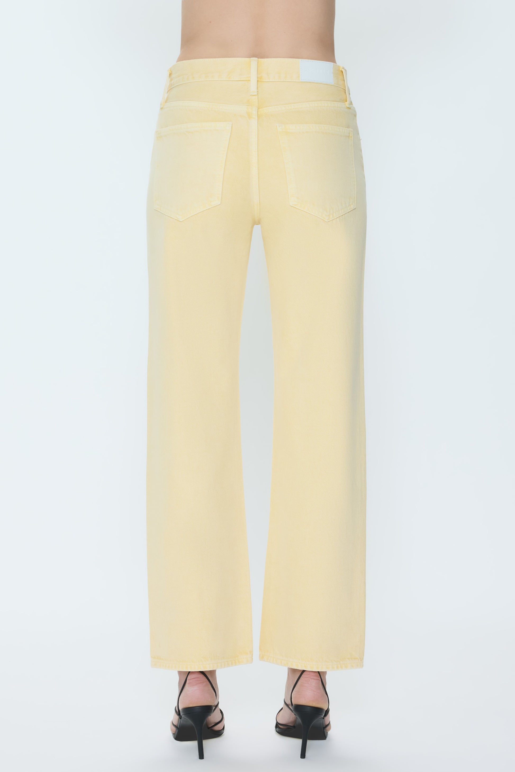 A person is shown from the back against a white background wearing Pistola's Lexi - Mid Rise Bowed Straight jeans in light yellow and black heeled sandals.