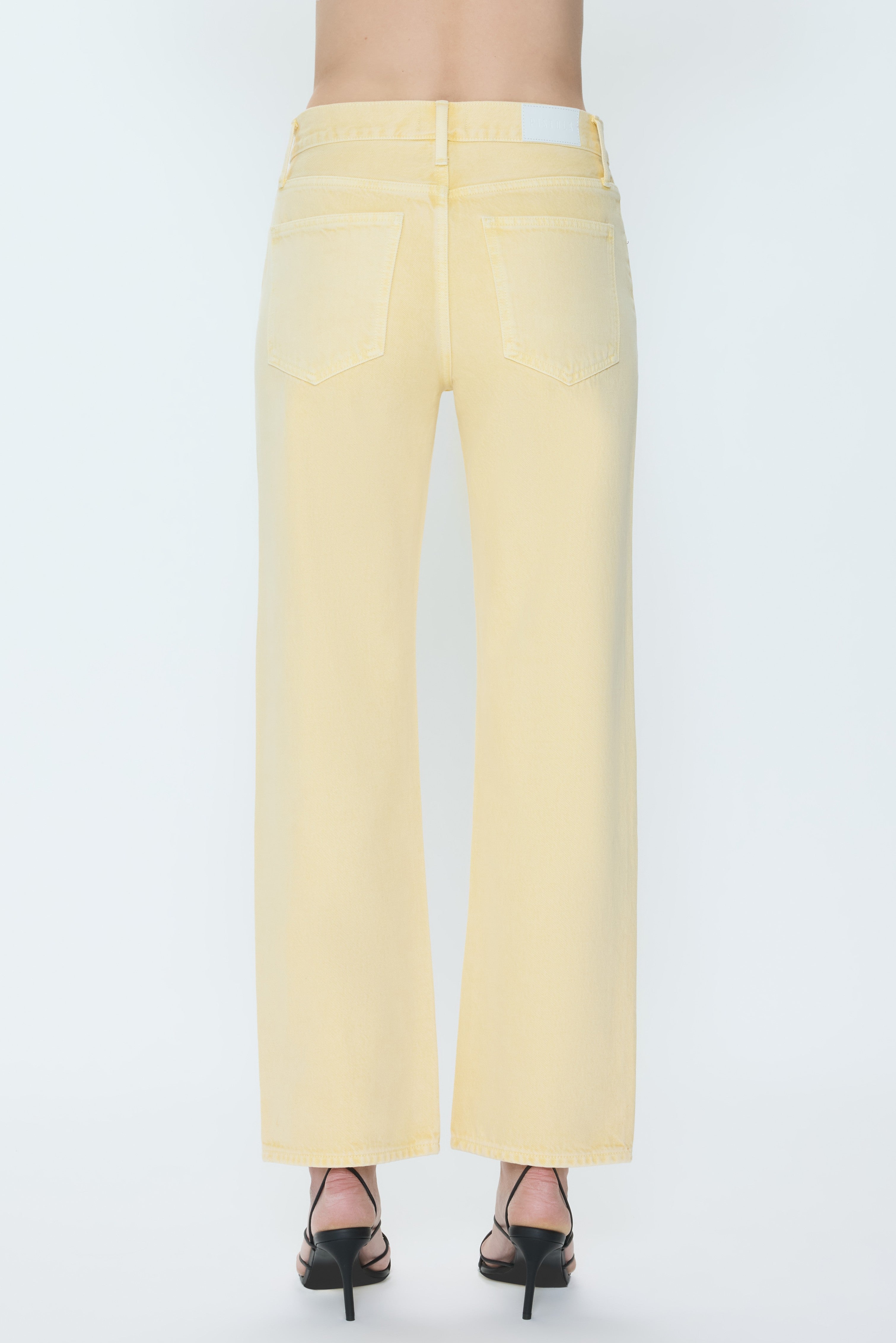 A person is shown from the back against a white background wearing Pistola's Lexi - Mid Rise Bowed Straight jeans in light yellow and black heeled sandals.