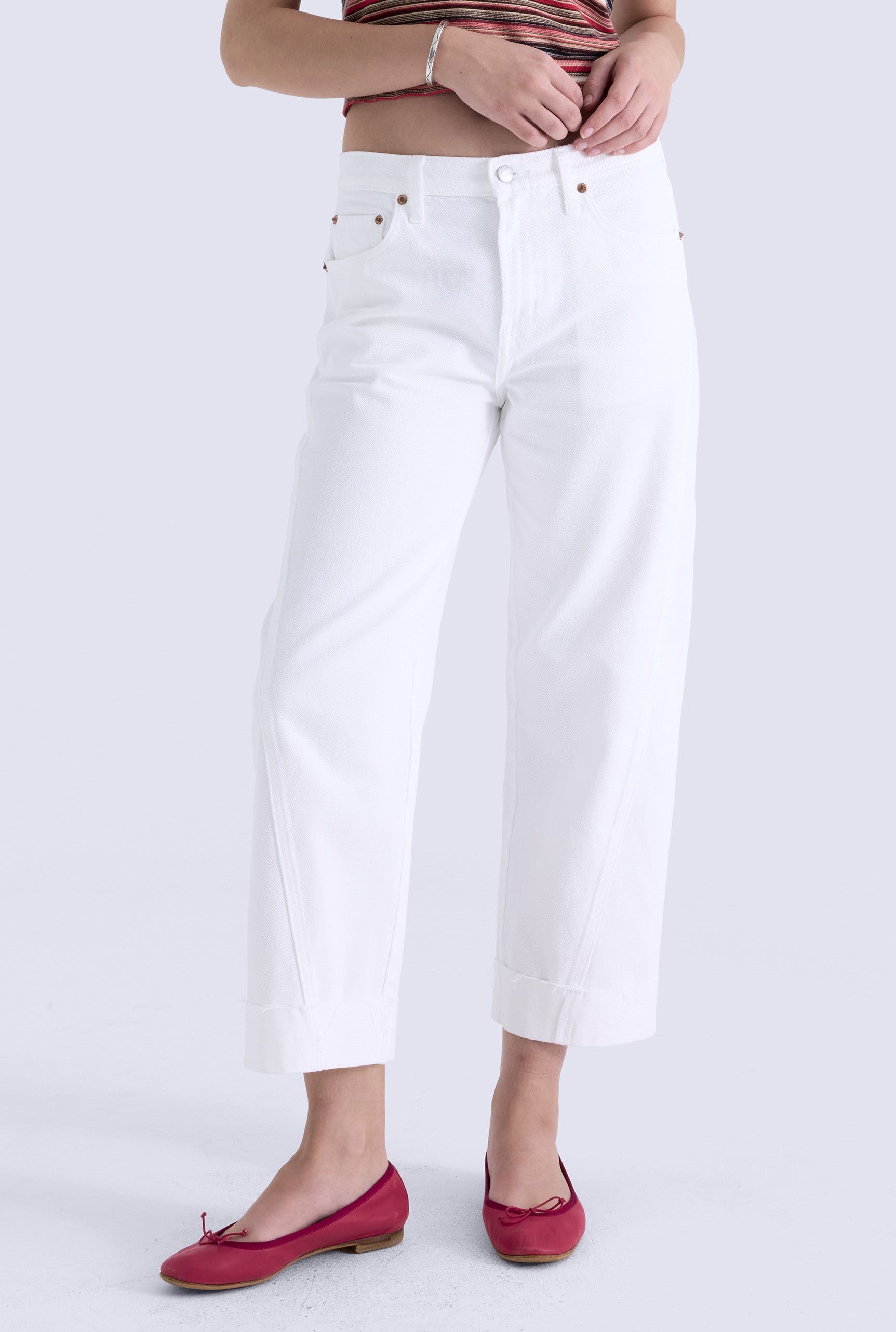 A person wears Pistola's Kacey Mid Rise Cuffed Boyfriend jeans in white, paired with a striped crop top and red ballet flats, standing against a light background. Only the body from the shoulders down is visible.