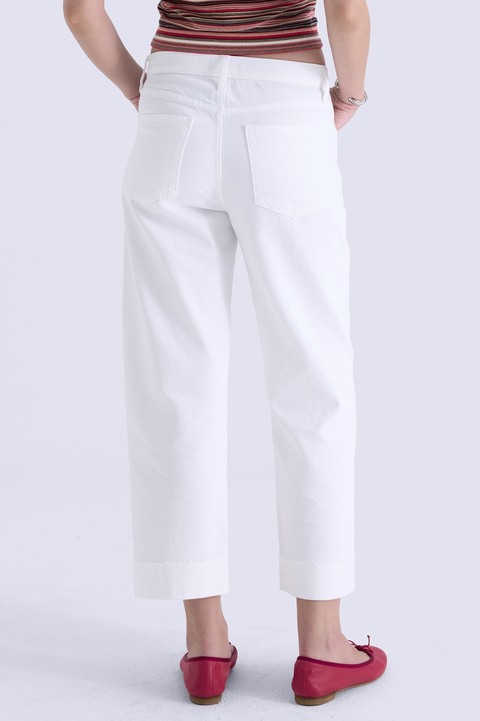 Facing away, a person wears Pistola’s Kacey Mid Rise Cuffed Boyfriend jeans in white rigid denim with two large back pockets, paired with a sleeveless striped top and red ballet flats. The jeans have a cuffed hem above the ankle. Background: plain light gray.