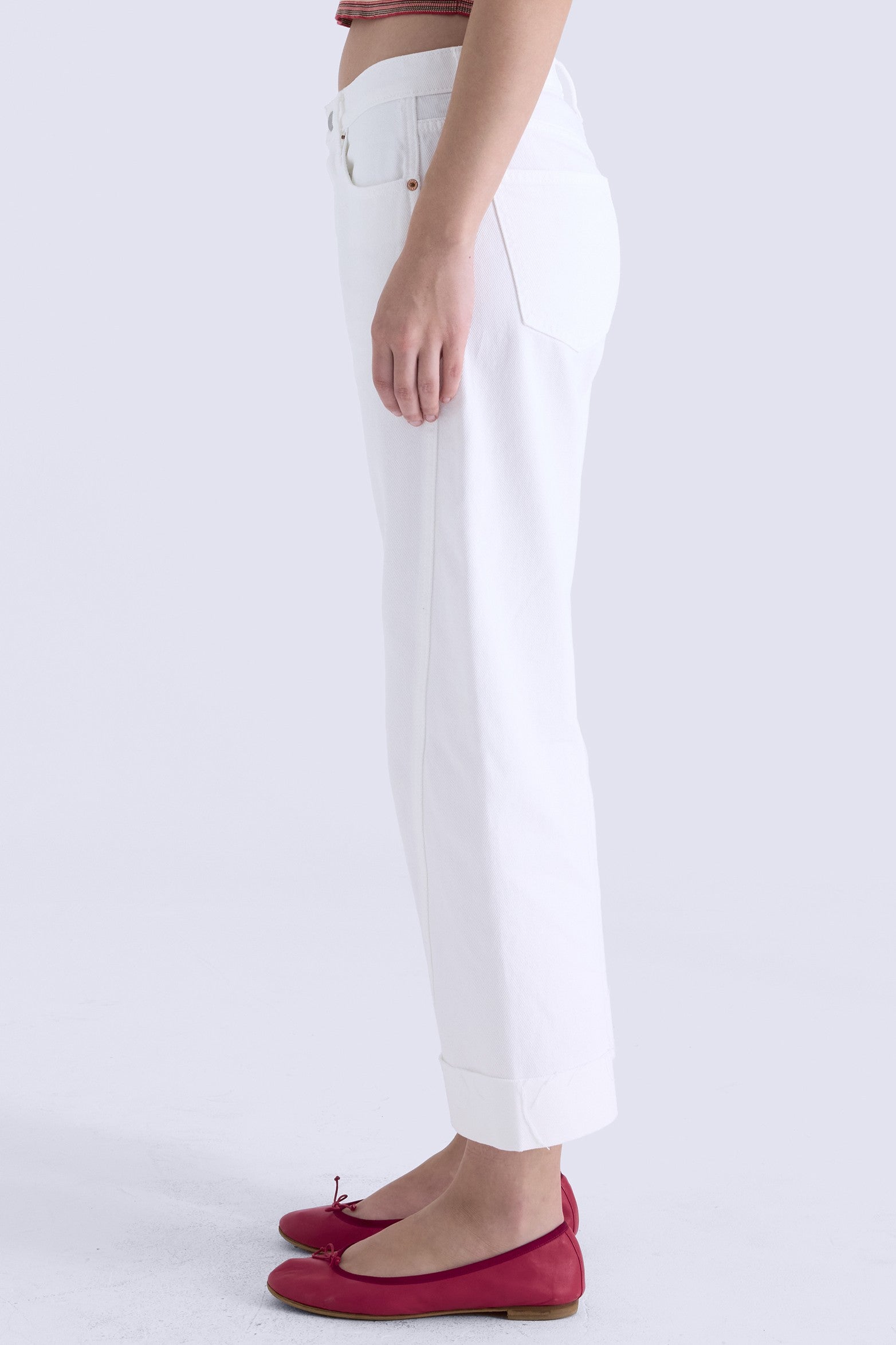 A person stands sideways against a plain light background wearing Pistola's Kacey - Mid Rise Cuffed Boyfriend jeans in white, paired with red flats and their arm relaxed by their side.