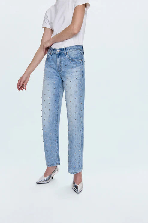 Wearing Pistola's Lexi In Confetti light blue high-waisted jeans with stud embellishments, a white short-sleeve top, and shiny silver pointed-toe heels, the person stands against a plain light background.