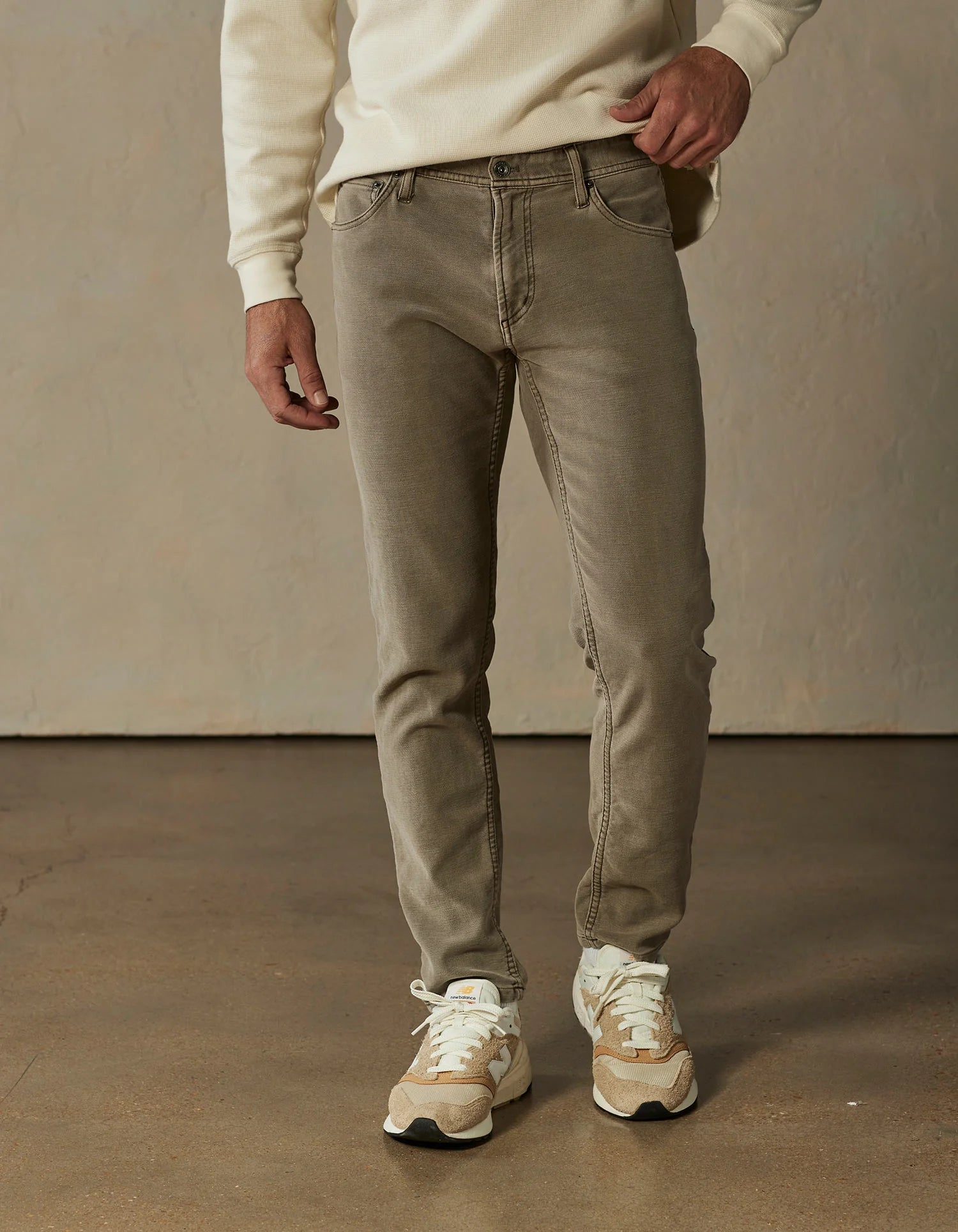 A person wears The Normal Brand Comfort Terry Pant in taupe, paired with a cream sweater and beige sneakers, standing indoors on a polished brown floor against a neutral wall. Their face is not visible.