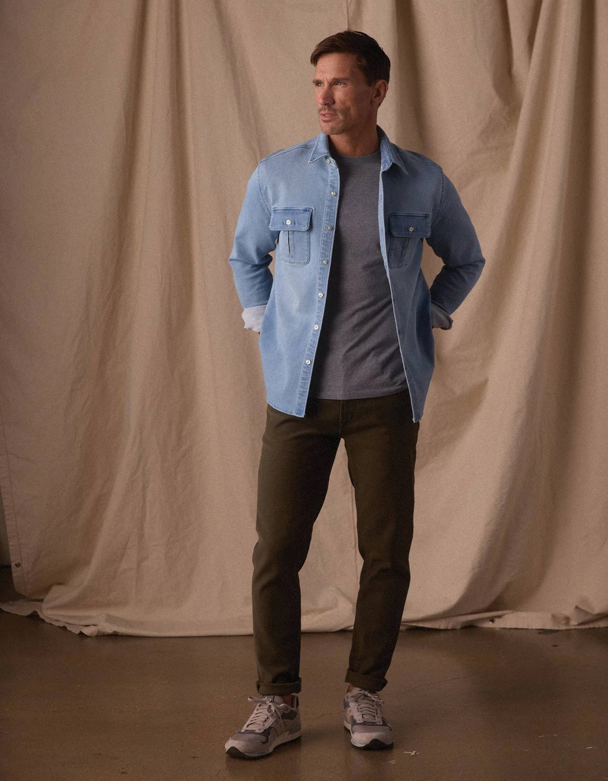 A man stands indoors against a beige fabric backdrop, wearing a light blue button-up over a gray tee, The Normal Brand Comfort Terry Pant in Olive, and gray sneakers, with his hands in his pockets and looking to the side.