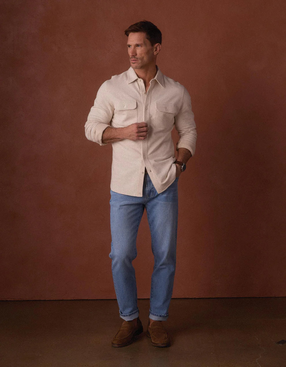 A man stands against a brown background wearing The Normal Brand's Textured Knit Shirt, light blue jeans, and brown shoes, with one hand in his pocket and the other resting on his thigh.