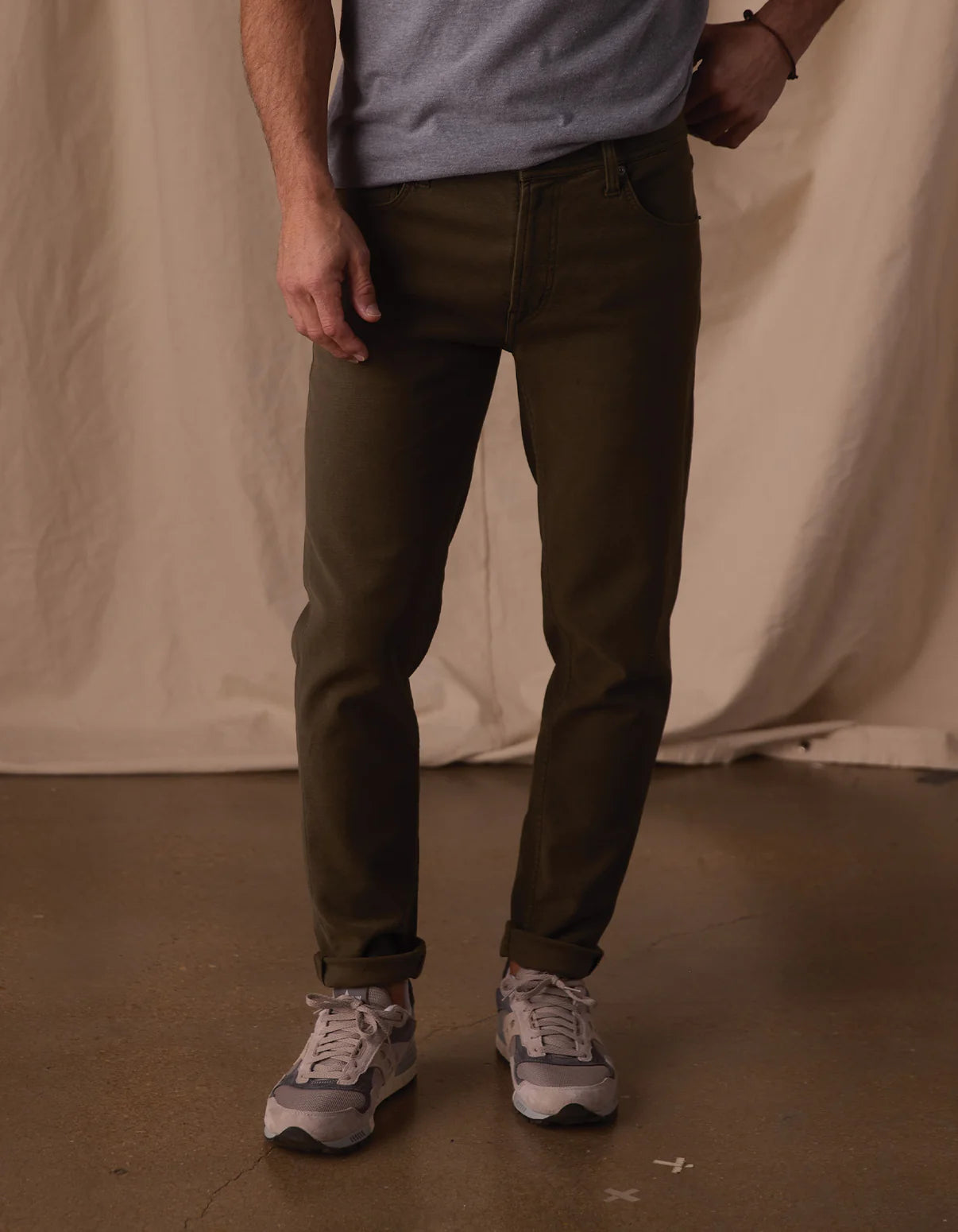 Only the lower half of a person is visible, wearing The Normal Brand's Comfort Terry Pant in Olive, paired with a light gray t-shirt and gray sneakers. They stand on a brown floor against a beige fabric backdrop.