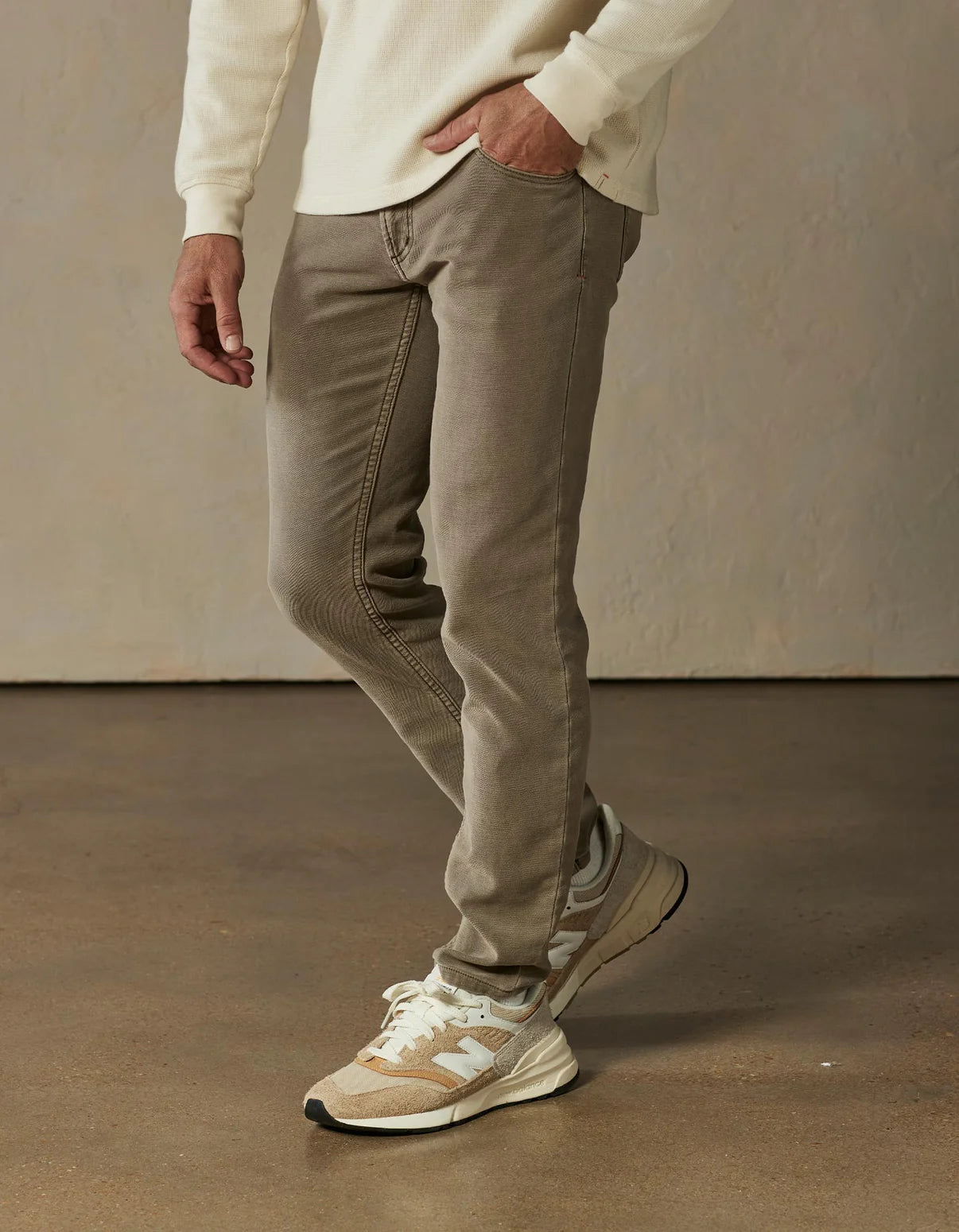 Wearing The Normal Brand Comfort Terry Pant in Taupe and a cream long-sleeve shirt with beige New Balance sneakers, a person stands on polished concrete against a light textured wall.
