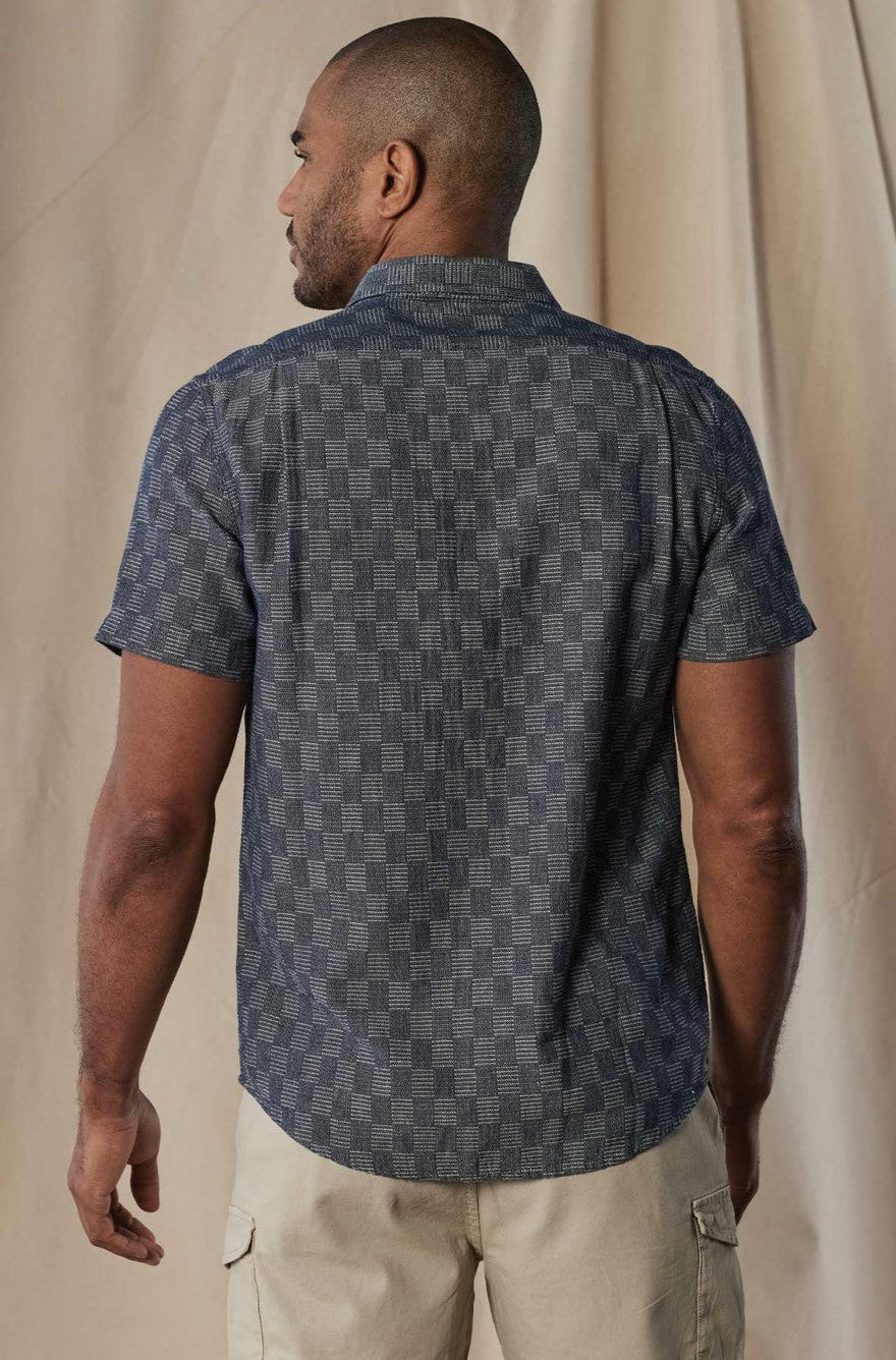 A man, back turned, wears The Normal Brand’s Freshwater Button Up Ivory Crinkle shirt with short sleeves and shirttail hem, paired with light shorts, standing before a beige, slightly wrinkled fabric backdrop.