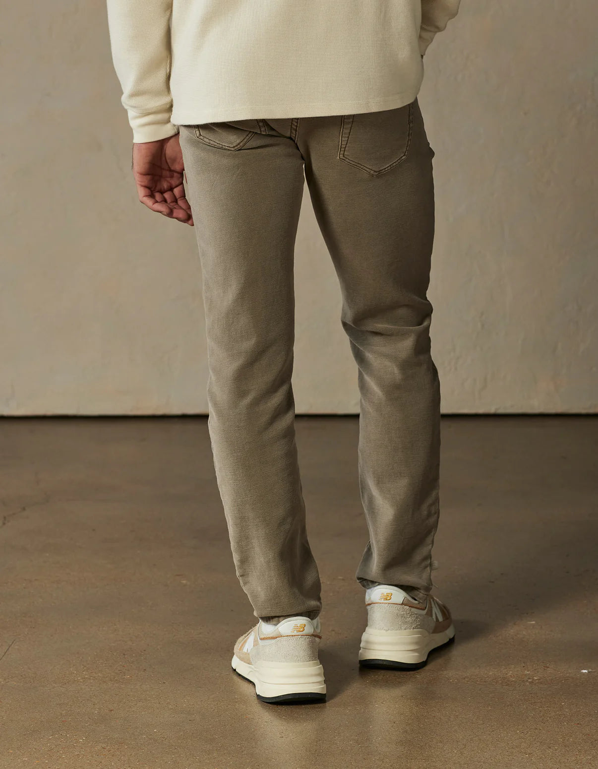 A person stands indoors with their back to the camera, wearing The Normal Brand’s Comfort Terry Pant in Taupe, a cream long-sleeve shirt, and white sneakers on a polished concrete floor.