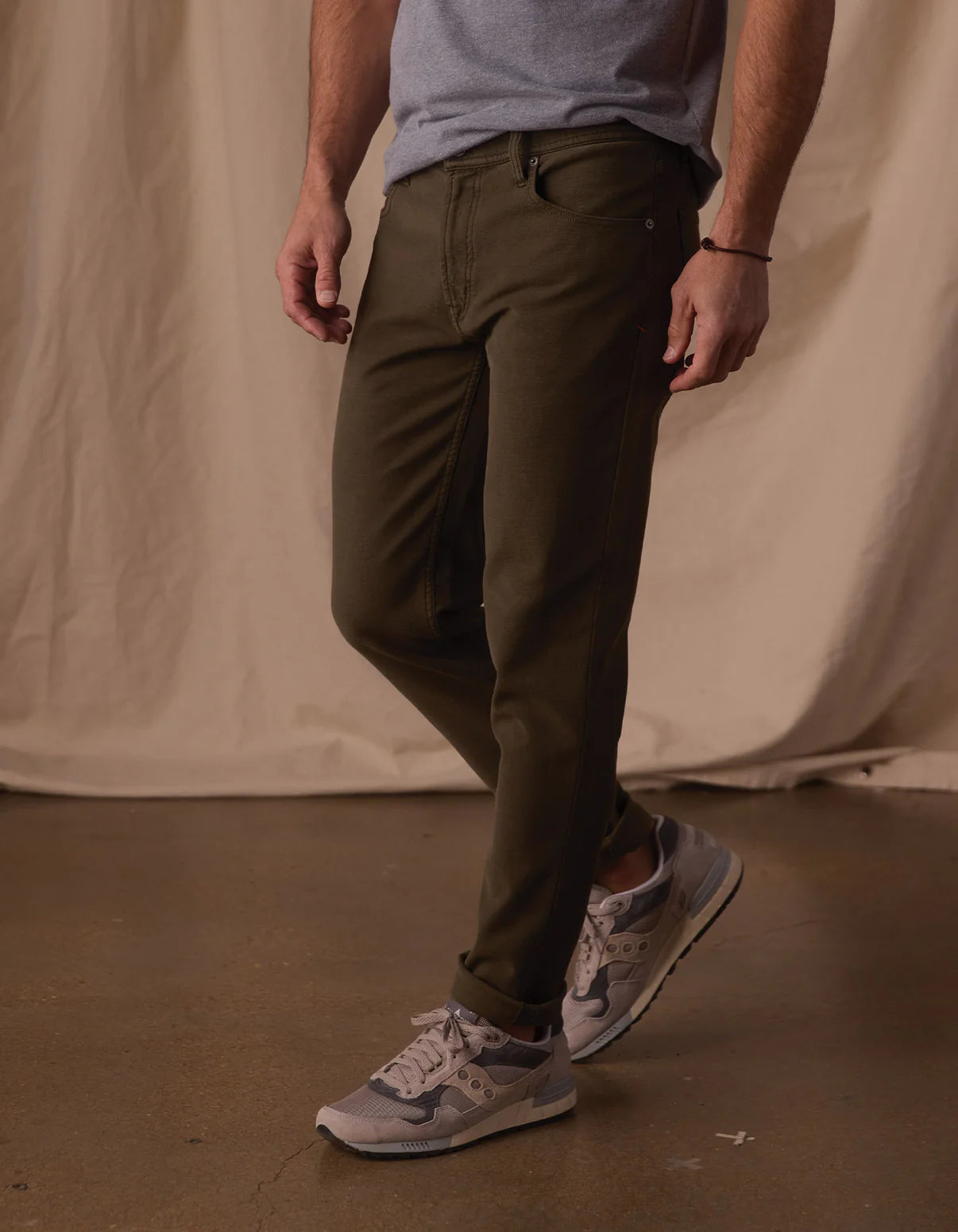 A person wearing The Normal Brand Comfort Terry Pant in Olive, a light gray t-shirt, and gray sneakers stands on a polished floor before a beige fabric backdrop. The person’s face is not visible.