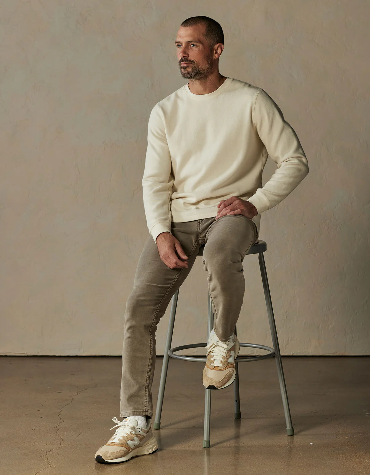 A man with short hair and a beard sits on a stool in a minimalist, neutral-toned setting, wearing The Normal Brand’s Comfort Terry Pant in Taupe, paired with a cream sweater and beige sneakers.