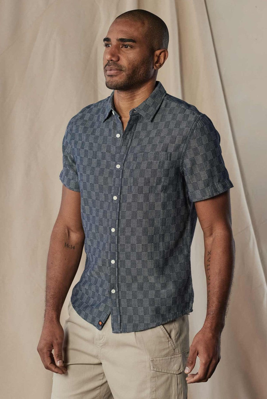 A man wears The Normal Brand's Freshwater Button Up Ivory Crinkle, a short-sleeved, button-up shirt, paired with light pants and standing against a neutral draped fabric background.