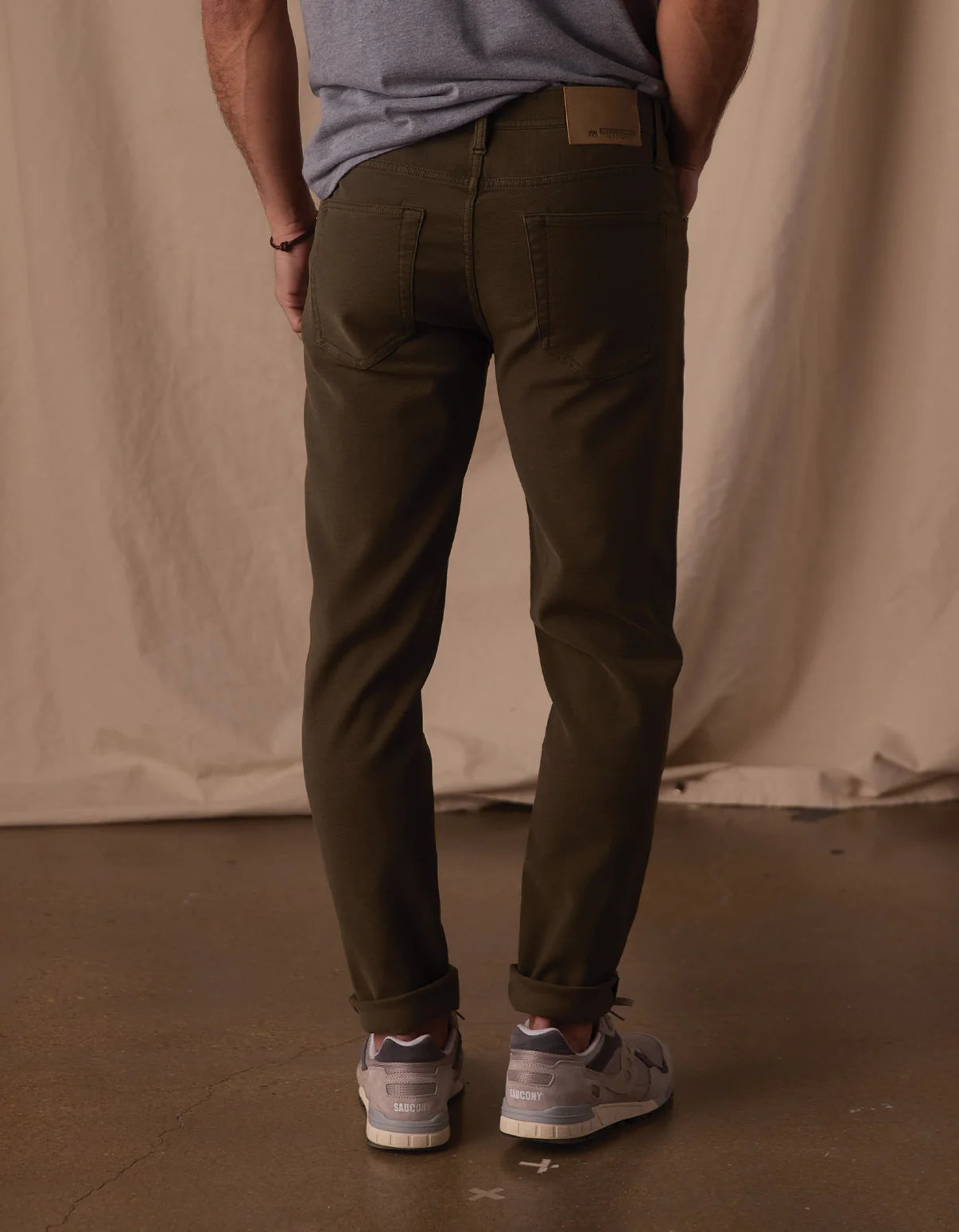 A person stands indoors facing away, wearing The Normal Brand's Comfort Terry Pant in Olive with a gray T-shirt and sneakers, against a backdrop of beige fabric drape and polished concrete floor.