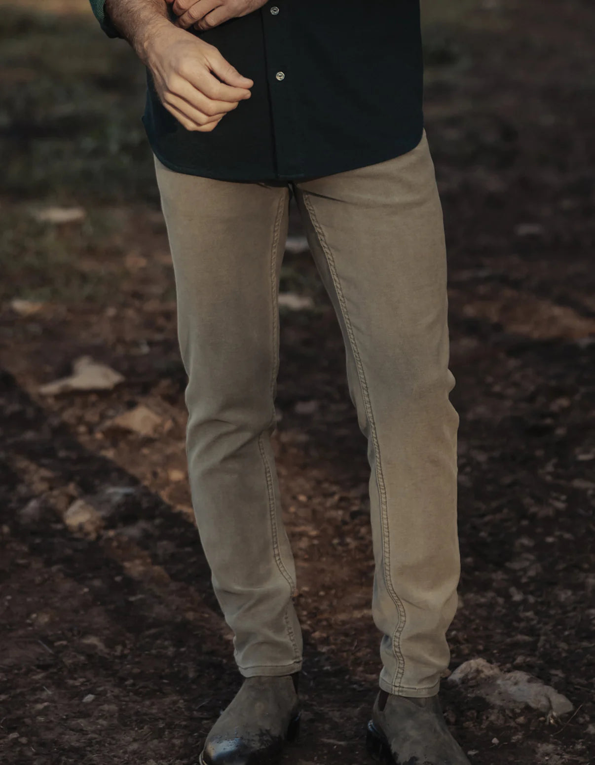A person wears The Normal Brand Comfort Terry Pant in Olive with dark boots and a long-sleeve shirt, standing on a dirt path with grass and rocks, only their lower body visible.