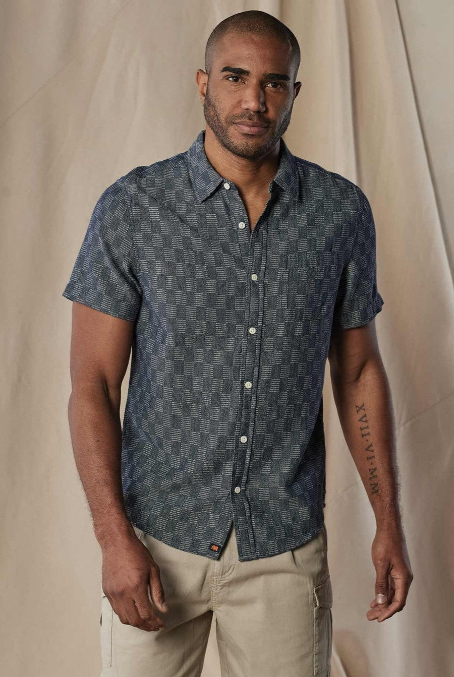 A man with closely shaved hair and a trimmed beard wears The Normal Brand’s Freshwater Button Up Ivory Crinkle in Navy Multi, paired with beige cargo pants. He stands against a neutral backdrop, his arm tattoo partially visible.