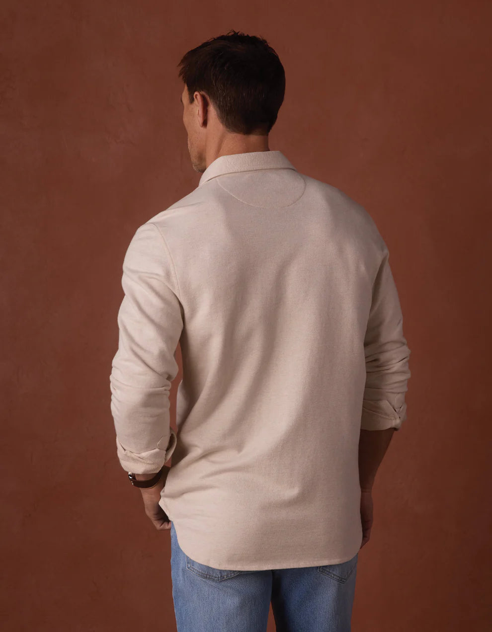A man stands with his back to the camera, wearing The Normal Brand’s Textured Knit Shirt in light beige and light blue jeans, set against a plain brown background.
