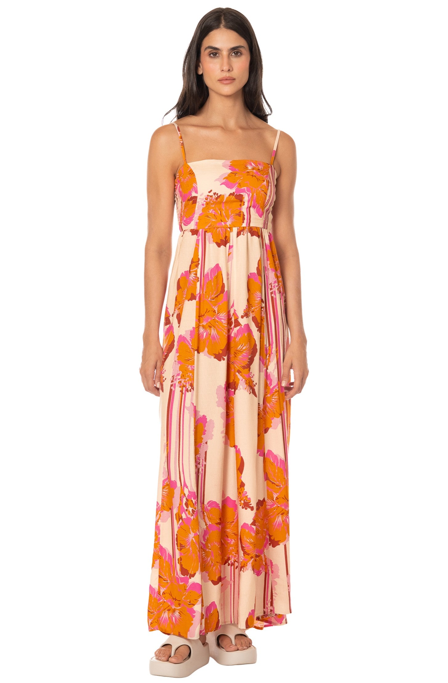 A woman wears the Maaji Molokai Bloom Ela Dress in Orange Multi, a sleeveless maxi with adjustable straps and lace-up back, featuring vibrant floral patterns. She stands against a white background with light-colored sandals.