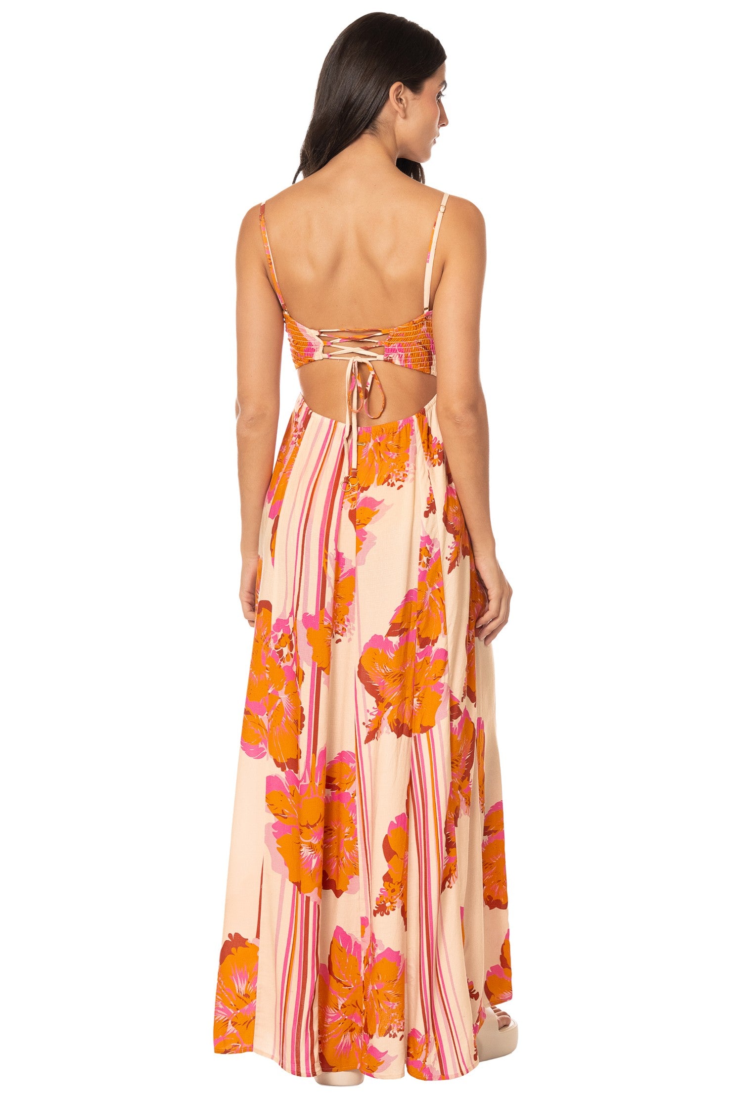 A woman faces away from the camera in the Maaji Molokai Bloom Ela Dress—a long, flowy maxi with orange floral and pink stripes, adjustable straps, an open lace-up back, and a center tie detail.