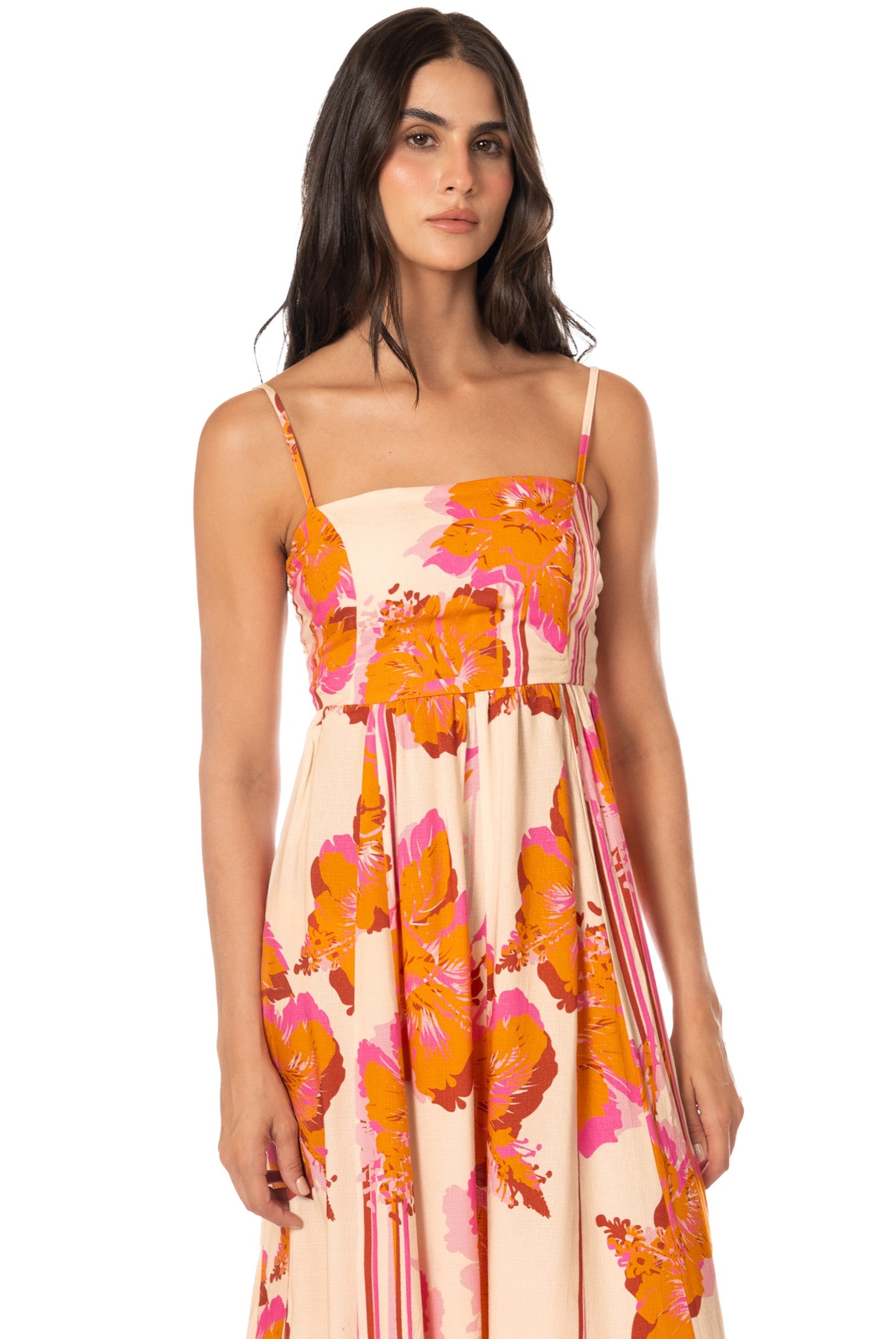 A woman with long dark hair wears the Maaji Molokai Bloom Ela Dress, a sleeveless, floral-print maxi in pink and orange with adjustable straps, standing against a plain white background.