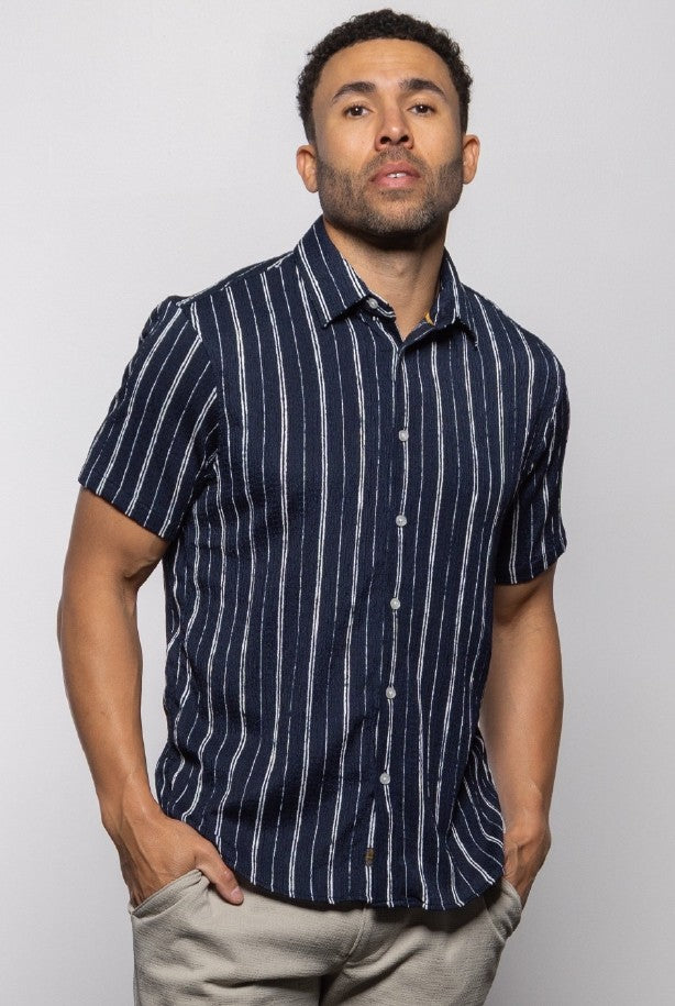 A man confidently poses in a Mason Cruz Bali Short Sleeve Shirt—relaxed-fit, navy with white stripes—paired with light pants, perfect for warm weather against a plain background.