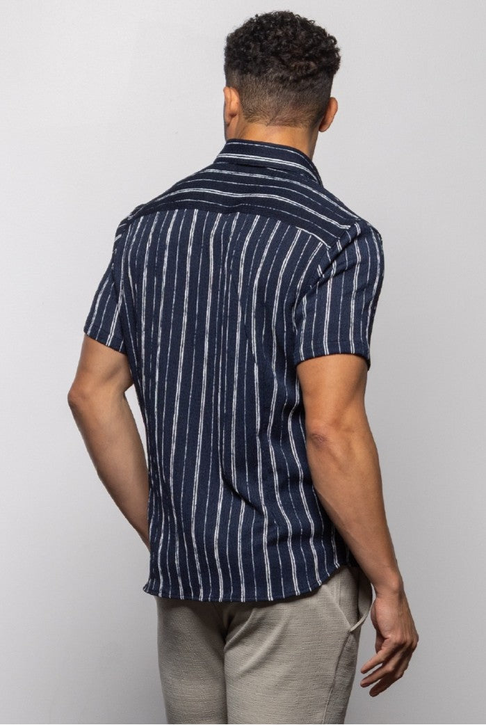 A man faces away from the camera in Mason Cruz's Bali Short Sleeve Shirt, a navy blue and white striped relaxed-fit piece, paired with light beige pants—perfect for warm weather—set against a plain light gray background.