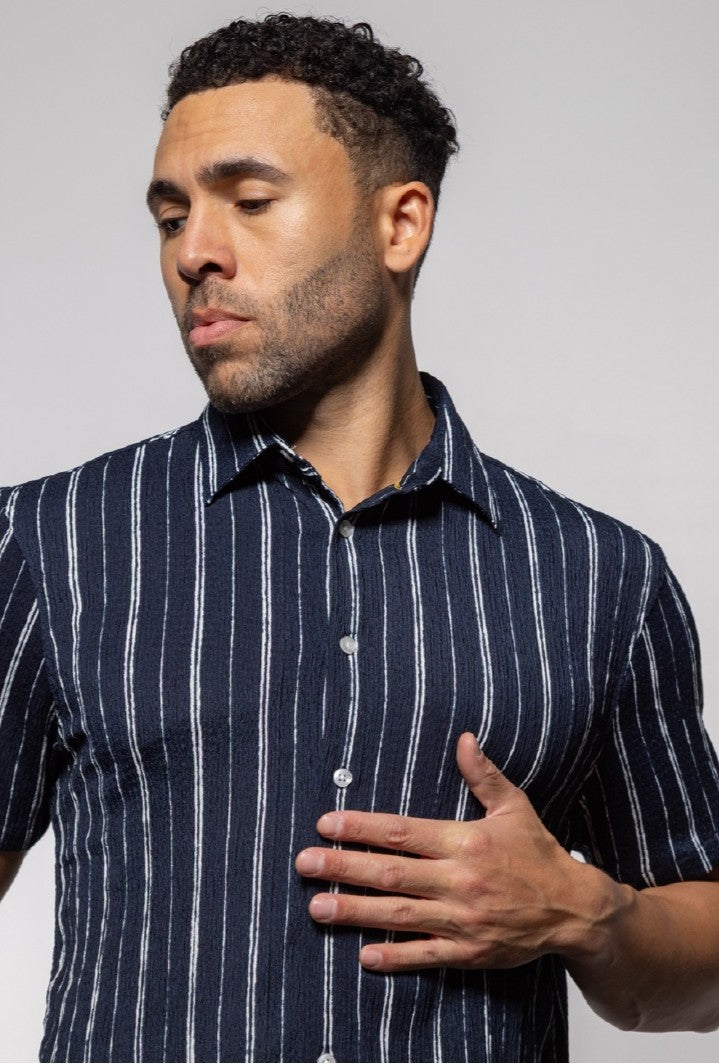 A man wears the Mason Cruz Bali Short Sleeve Shirt, a navy blue relaxed-fit shirt with white stripes, as he looks to his left and rests his hand on his chest against a plain gray background—ideal for warm weather.